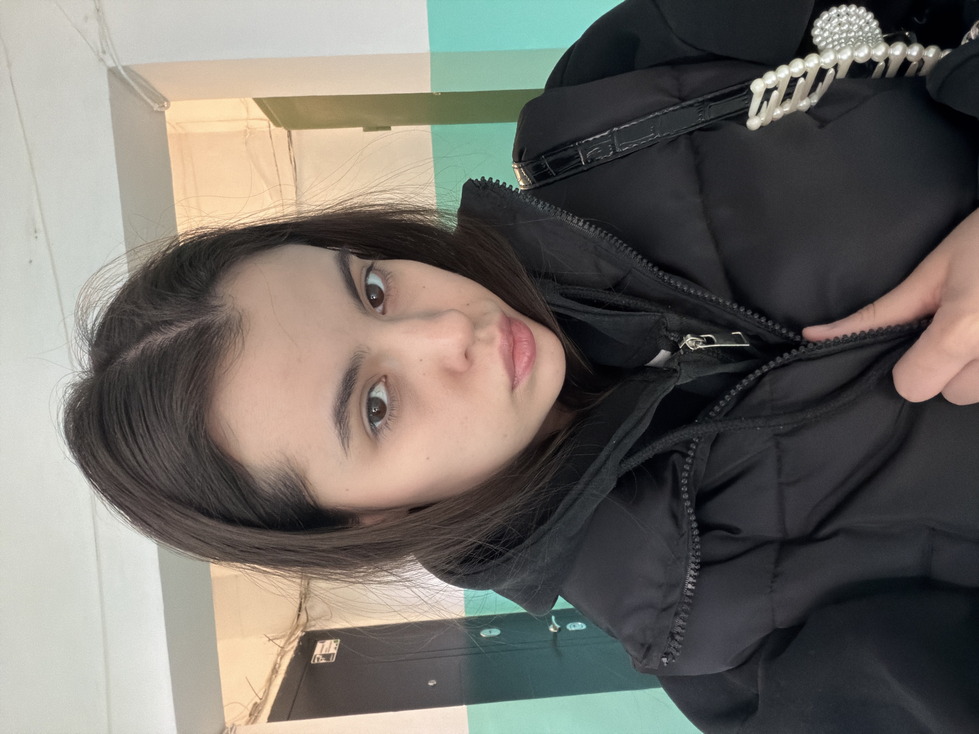 A young woman is centered in a vertical, eye-level shot. Her face is slightly angled towards the viewer. She has dark hair and brown eyes and wears a black puffer jacket. A black purse with a pearl-like clip is visible on her shoulder. The woman appears to be closing the zipper of her jacket with her hand.
The background shows a corridor with walls painted in alternating horizontal stripes of white, green, and light blue. A closed door is partially visible to the left. The location is assumed to be in the city of Chaikovsky, Russia. The lighting in the corridor is bright and somewhat sterile. Electrical wiring is visible along the top of the wall.