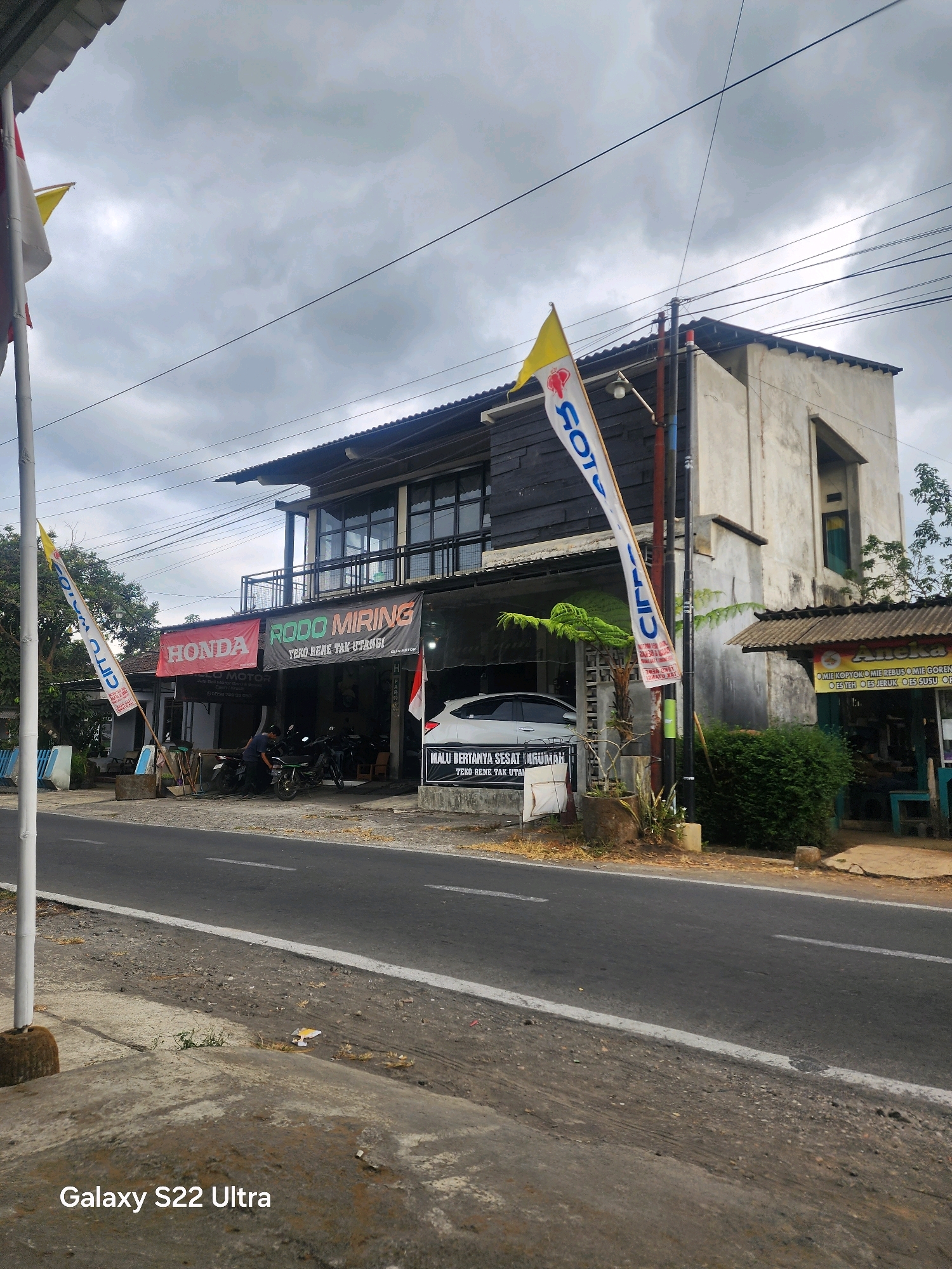 This is a street-level, medium-angle, vertical photograph taken on a cloudy day in Salaman, Indonesia. The scene depicts a row of shops and a street.
In the foreground, a paved road with a white line marking runs horizontally across the image. To the left of the road, a concrete sidewalk is visible with some dry grass and debris. A white pole with a brown base stands on the sidewalk. The bottom left corner of the image shows a watermark reading "Galaxy S22 Ultra."
The middle ground features a building with multiple storefronts. The leftmost establishment has a red banner with "HONDA" in white text and a smaller banner below that reads "RODO MOTOR". To the right of this is a shop with a dark grey facade and a black banner that says "RODO MIRING" in white and red text, with the tagline "TEKO RENE TAK URUS" below it. A white flag with blue text, partially obscured by a yellow and white flag, hangs from a pole in front of this shop. A white car is parked in front of this building. To the right, another shop has a small awning with colorful text advertising food items. A green bush and a potted plant are situated in front of this shop. Two individuals, appearing to be men, are standing near motorcycles outside one of the shops.
The background shows a multi-story building with a dark upper section and a light-colored, textured lower section. Windows are visible on the upper floor. A cluster of power lines crisscrosses the sky above the buildings. The sky is overcast with thick grey clouds. Trees and foliage are visible to the right of the buildings.