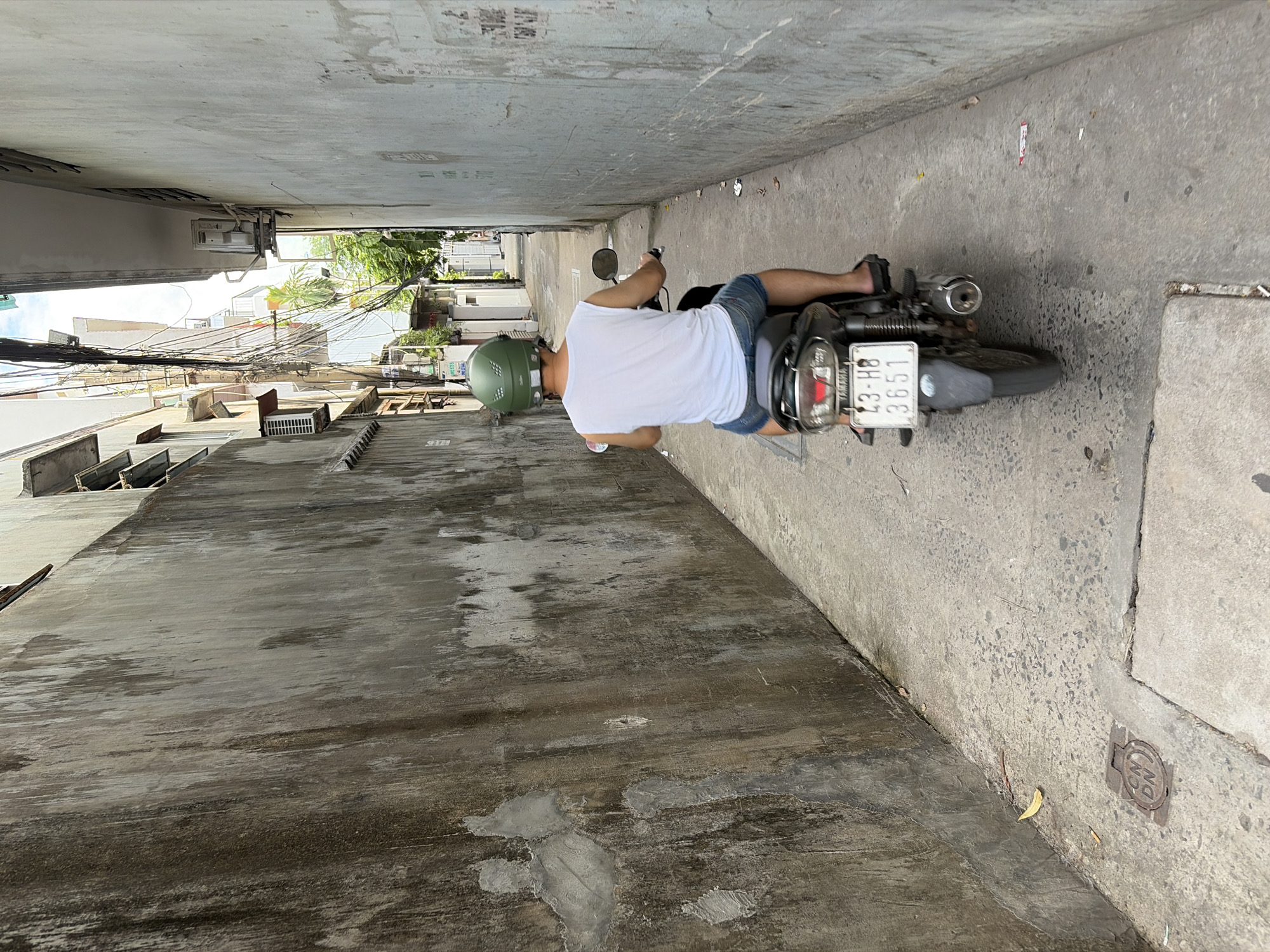 A person wearing a white tank top and denim shorts is riding a dark-colored motorcycle down a narrow alleyway in Da Nang, Vietnam. The rider has a green helmet on. The alley is flanked by tall, weathered concrete walls on both sides. The wall on the left is significantly darker and more textured, showing signs of water stains and patches of repair. The wall on the right is lighter in color and appears smoother, with some faint markings. The alley floor is paved with concrete and shows cracks and some scattered debris, including leaves and small pieces of paper. In the distance, the alley opens up slightly, revealing more buildings, electrical wires, and trees. A license plate on the back of the motorcycle reads "23-HB 3651". A metal grate cover is embedded in the concrete in the foreground of the left side of the alley.
