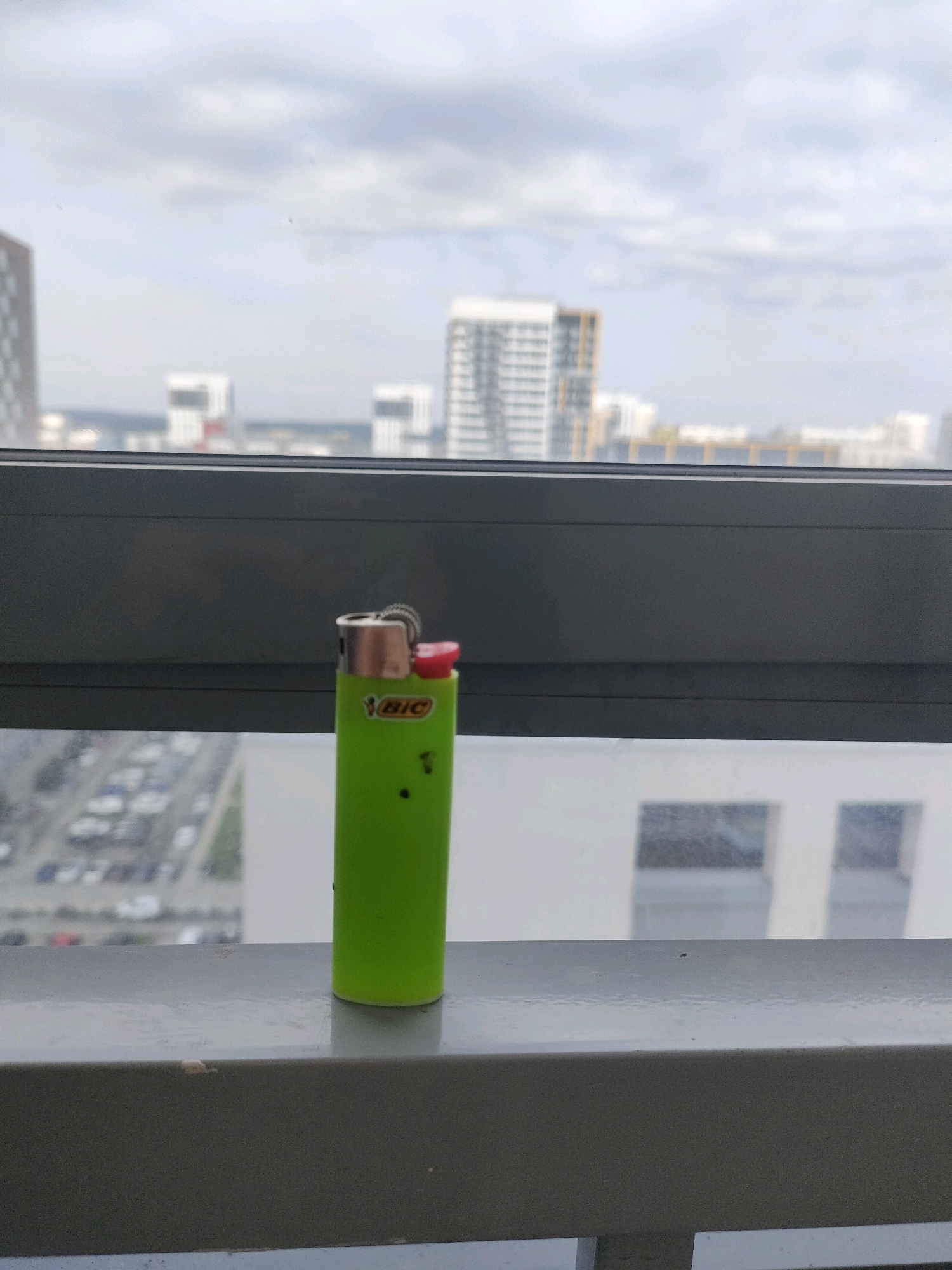 A bright green BIC lighter with a red button sits on a gray railing in front of a window. Through the window, a blurry cityscape of Yekaterinburg, Russia, is visible under a cloudy sky. The city features numerous modern buildings, some with distinctive architectural designs. In the foreground, a parking lot with many cars can be seen. The lighter is upright and centered in the frame, with its cap and ignition mechanism clearly visible. Two small dark specks are present on the surface of the lighter. The overall impression is one of an urban scene observed from a high vantage point.
