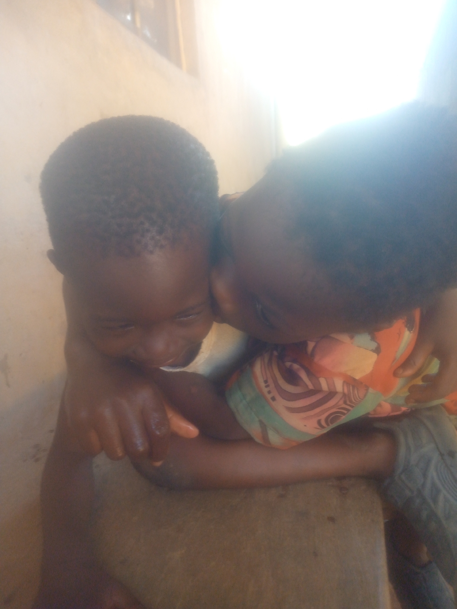 Two dark-skinned children are closely embracing each other. One child is wearing a colorful shirt with geometric patterns. They are positioned indoors, possibly sitting on a rough, stone-like surface. The location is unknown.