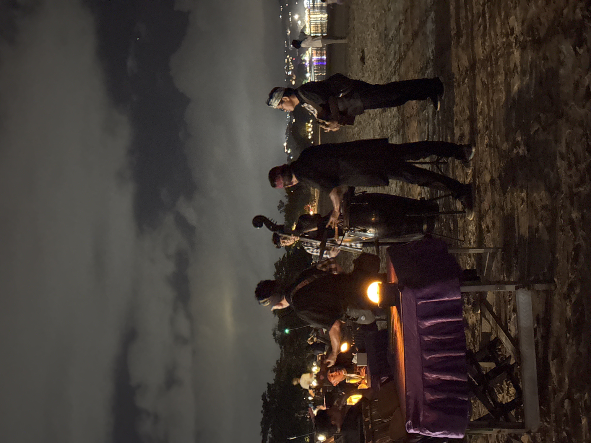 At night on a sandy beach, a group of musicians plays on a dimly lit stage. A double bass stands to the right, while two men in dark clothing are seen playing instruments. One is holding a guitar, and another appears to be drumming. In the foreground, a table covered with a purple cloth is lit by a bright lamp, and several people are seated around it. In the background, the city lights of Kuta Selatan, Indonesia, gleam across the water, and the sky is filled with clouds.