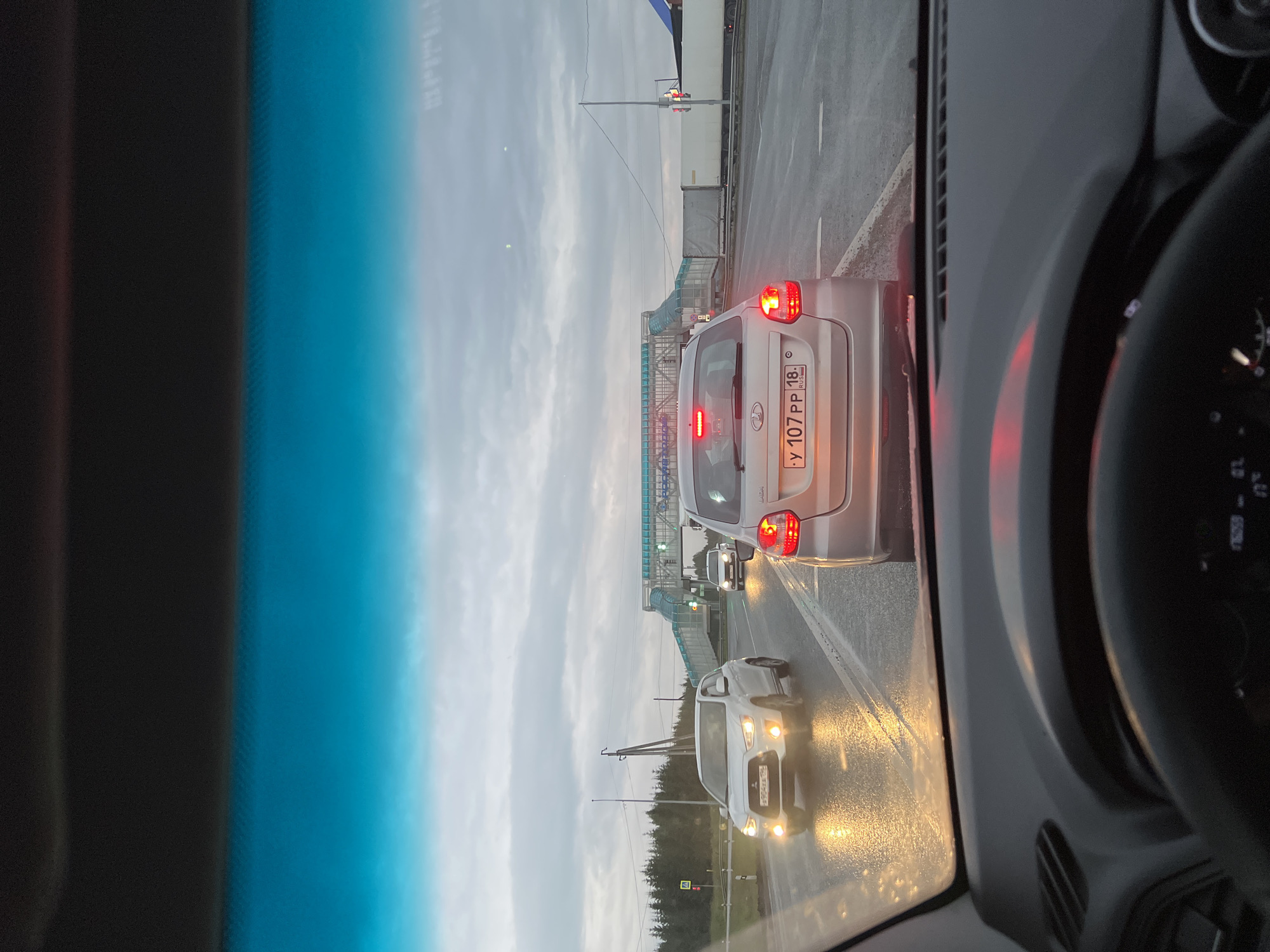 The image captures a view from inside a car, looking out at a traffic jam on a cloudy day, likely in the late afternoon or early evening, given the dim light. The road appears wet, suggesting recent rain. Several cars are visible, including a silver car in the foreground with its brake lights illuminated, and a white car further back with its headlights on. A pedestrian bridge spans the road in the distance. The scene is set on a multi-lane road, possibly a highway or major thoroughfare, with trees lining the sides. The sky is overcast with a mixture of grey and white clouds. The interior of the car is partially visible, including a dark-colored dashboard and steering wheel.
Visible text includes a license plate on the silver car which reads "У 107 РР 18 RUS". There is also some unreadable text on a screen or display within the car's interior on the right side of the image.