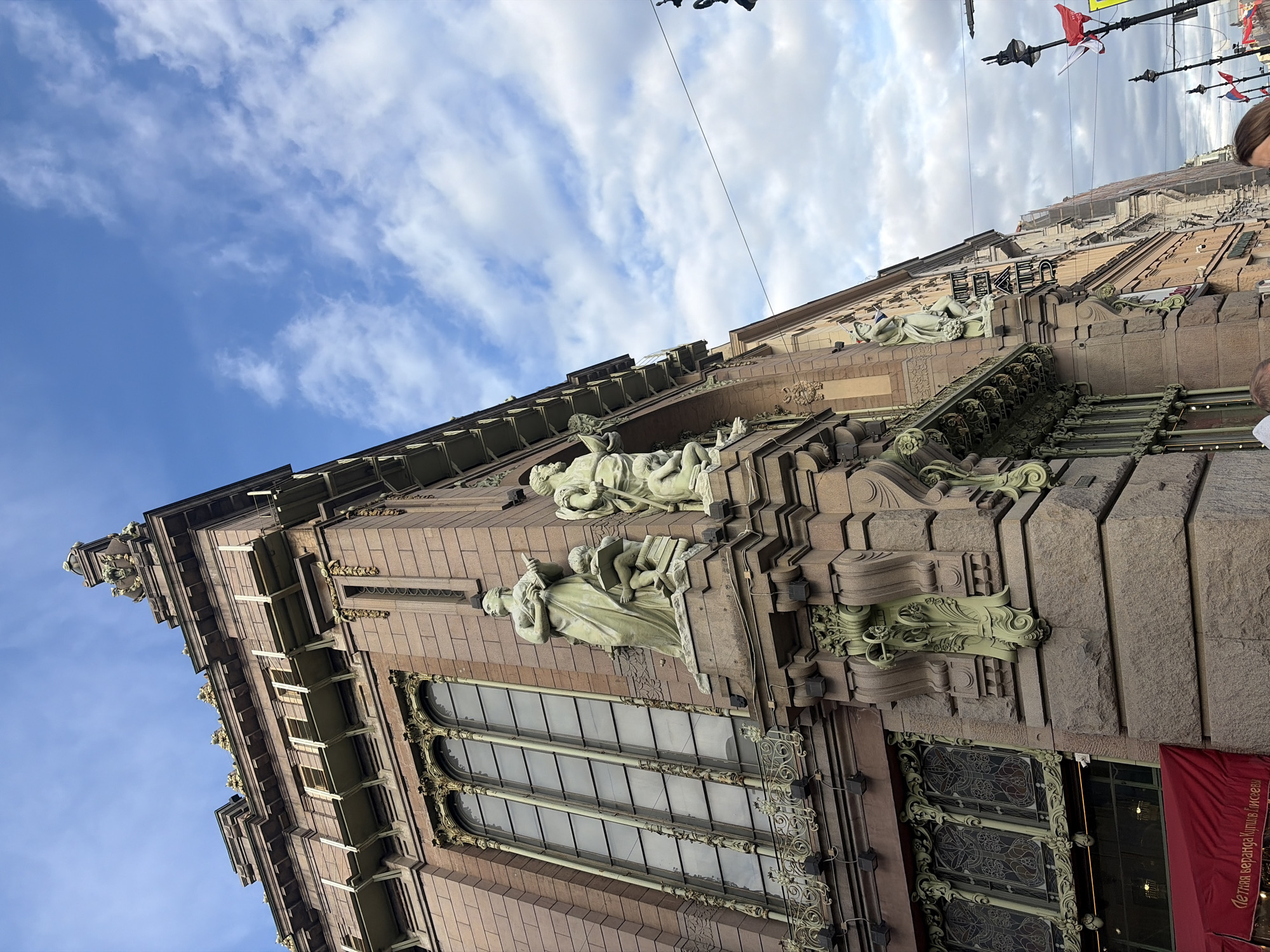 The image captures a section of the exterior of the Eliseyev Emporium, a historical landmark in Saint Petersburg, Russia. The building features Art Nouveau architecture with ornate sculptural elements and decorative ironwork. Two prominent statues are visible, depicting human figures, possibly representing arts or sciences, as commonly used in this architectural style. The building is made of stone in shades of brown and beige. It has large arched windows with intricate ironwork frames. Above the windows, there are decorative elements, including elaborate green sculptures. A sign with Cyrillic letters is also visible. In the background, the sky is partially clouded. There are power lines and flagpoles.