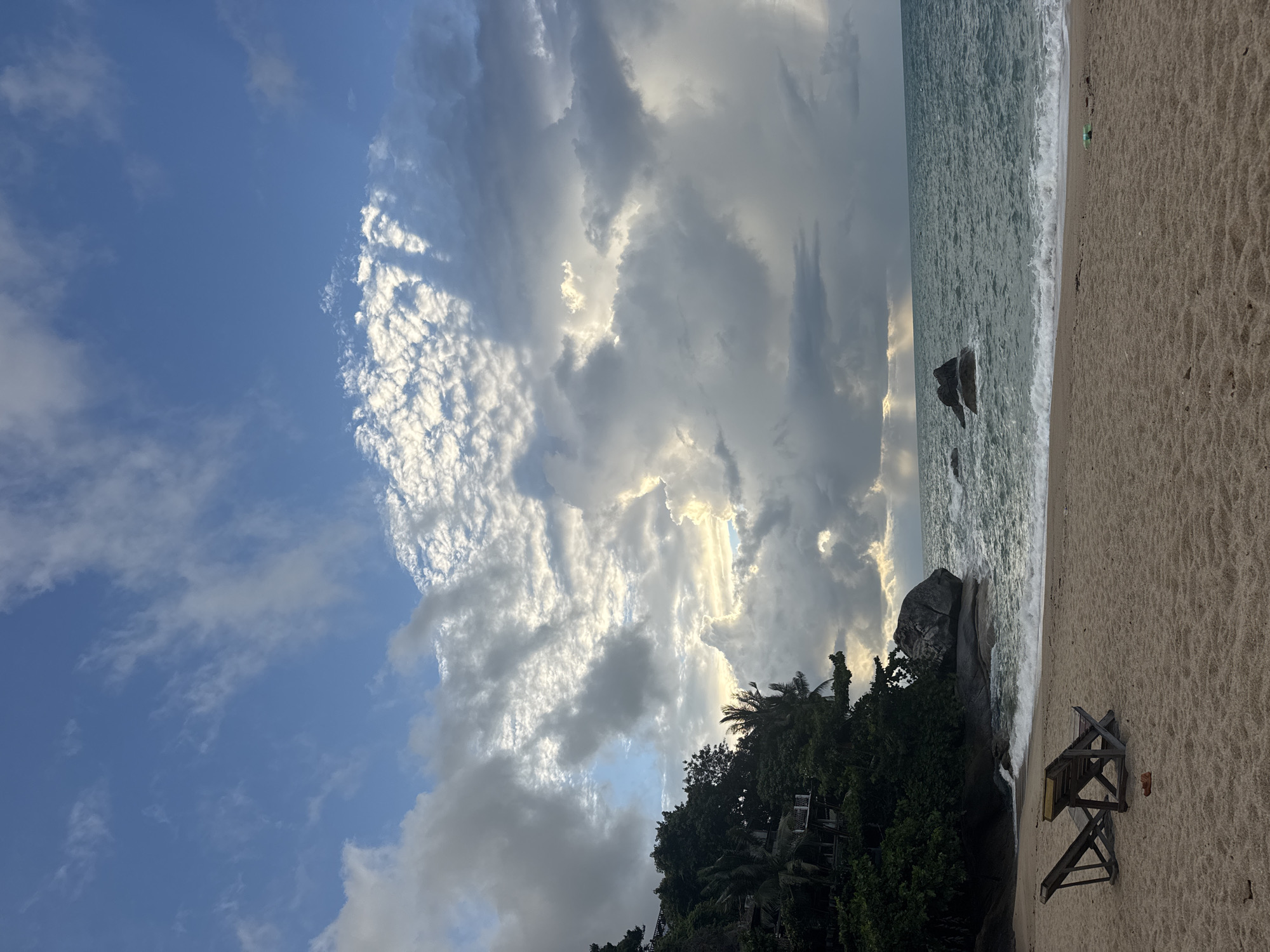 The scene is a beach in Baan Thong Nang, Thailand. The sky is partially cloudy with some blue sky visible, with the sun breaking through. The ocean meets a sandy beach with gentle waves. Some rocks and trees line the edge of the beach near the water. A beach chair is visible in the foreground.
