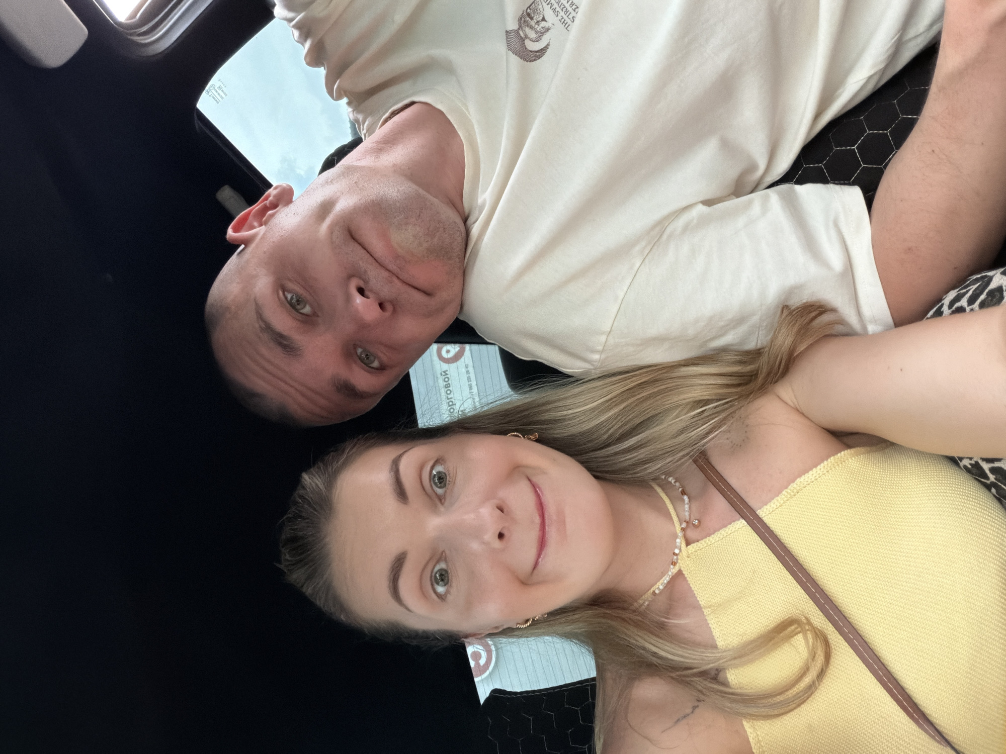 Two individuals, a woman and a man, are captured in a selfie taken from within a vehicle. The woman, positioned on the left, has long blonde hair and is looking directly at the camera with wide, surprised eyes and an open-mouthed smile. She is wearing a yellow top and a beaded necklace. The man, on the right, has a shaved head and is leaning his head against the woman's shoulder, looking towards the camera with a slight smirk. He is wearing a light-colored t-shirt. The background suggests the interior of a car, with a dark ceiling and a window showing an overcast sky. The location is identified as Gubarevo, Russia.