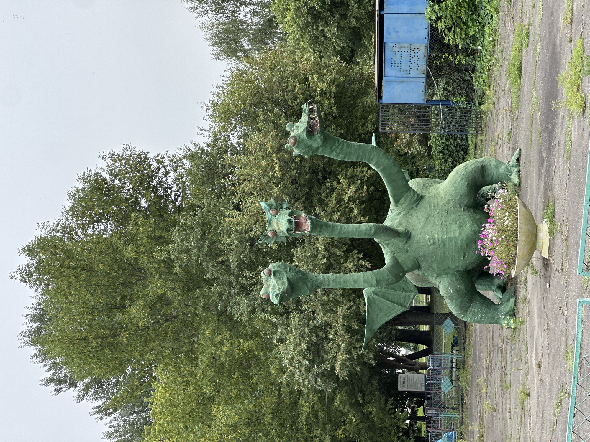 A three-headed green dragon sculpture is the main subject of the image, positioned in the foreground. The sculpture is detailed with wings, claws, and fangs. A planter with pink and white flowers sits in front of the dragon. The dragon is situated on a paved area surrounded by trees and foliage. In the background, there is a blue metal structure and fencing. The overall environment appears to be an outdoor space, possibly a park or an abandoned attraction, with a slightly overcast sky. The location is identified as the city of Biysk, Russia.