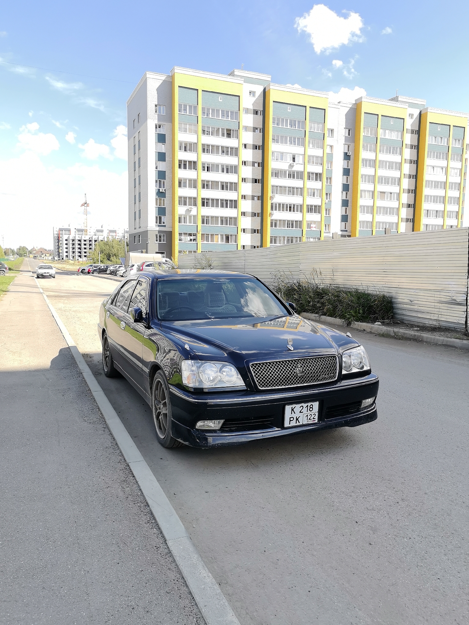 A black Toyota Crown sedan is parked on the right side of an asphalt road in Novoaltaysk, Russia, with its front facing the camera. The car has a chrome grille and a license plate reading "K 218 PK 122". In the background, a modern apartment building with yellow and grey facade elements stands under a partly cloudy blue sky. To the left of the car, the road curves towards the distance where other vehicles and unfinished construction projects are visible. A metal fence runs along the right side of the road behind the car. The scene appears to be a residential area under development.