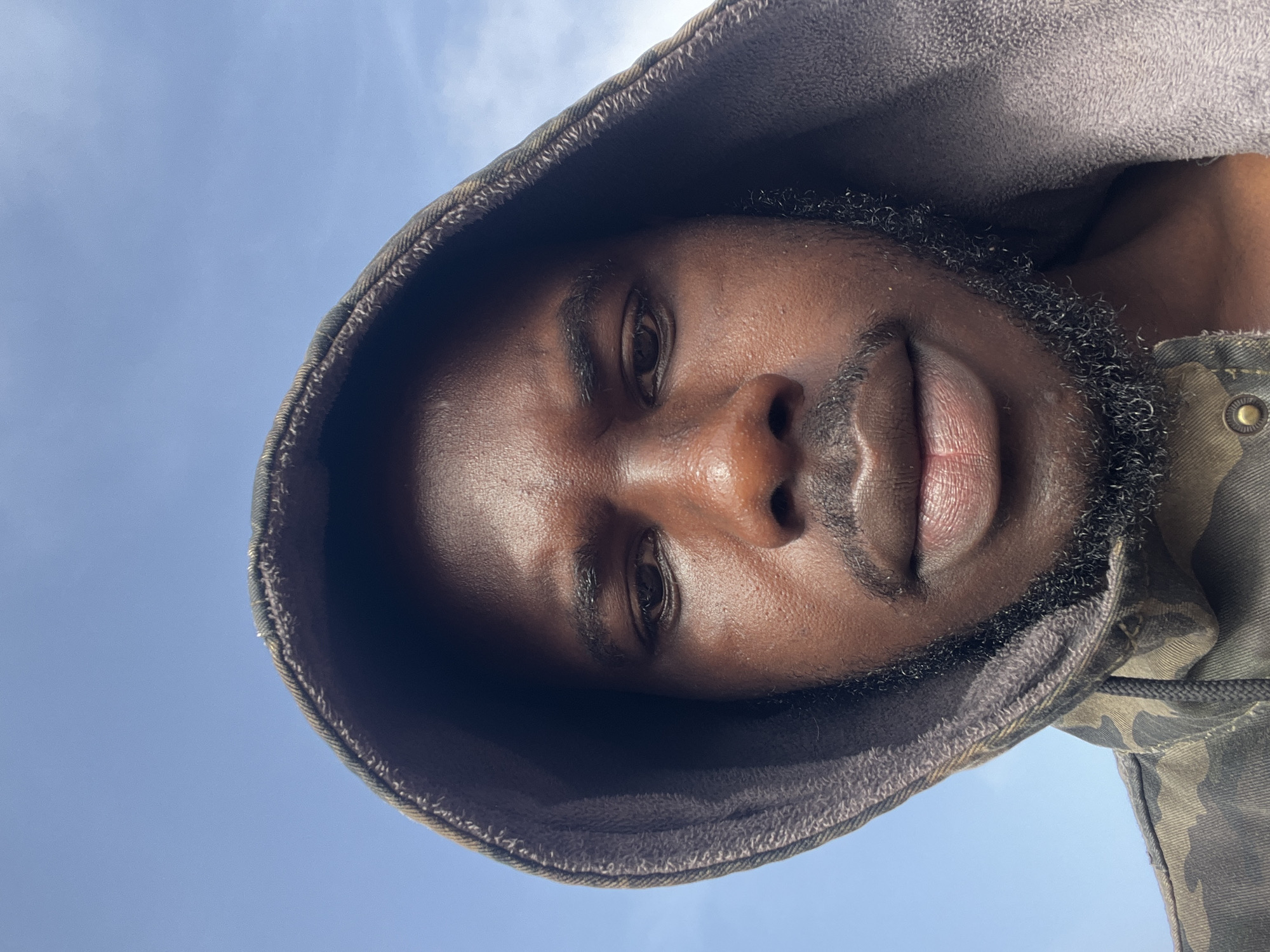 A close-up, upward-facing selfie of a Black man wearing a camouflage-patterned jacket with the hood pulled up. The man has dark skin, a beard, and is looking down. The background is a pale blue sky with some wispy clouds, suggesting an outdoor setting. The image appears to have been taken during the day. The specific location in Jos, Nigeria cannot be determined from the visual information provided in the image.