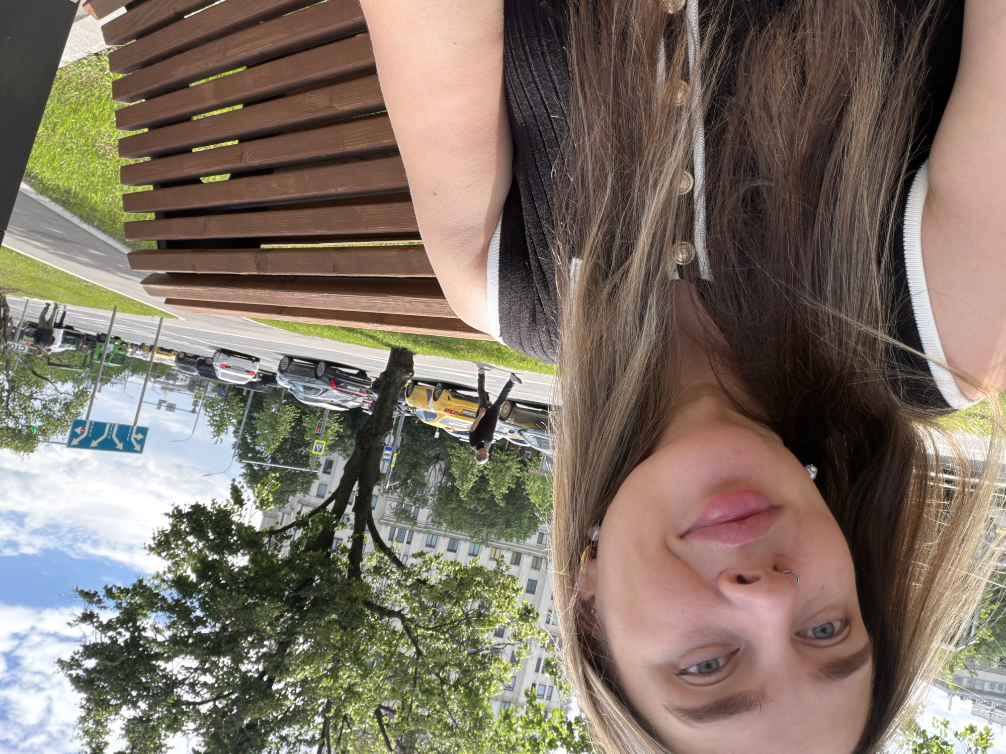 A selfie of a Caucasian female with long, light brown hair wearing a black and white top with gold buttons. She is sitting on a wooden bench in a park. The background shows a street with cars, buildings, trees, and a blue sky with clouds. A pedestrian is walking on the sidewalk. Traffic signs are visible. The location is likely Podolsk, Russia, given the urban setting and building architecture.