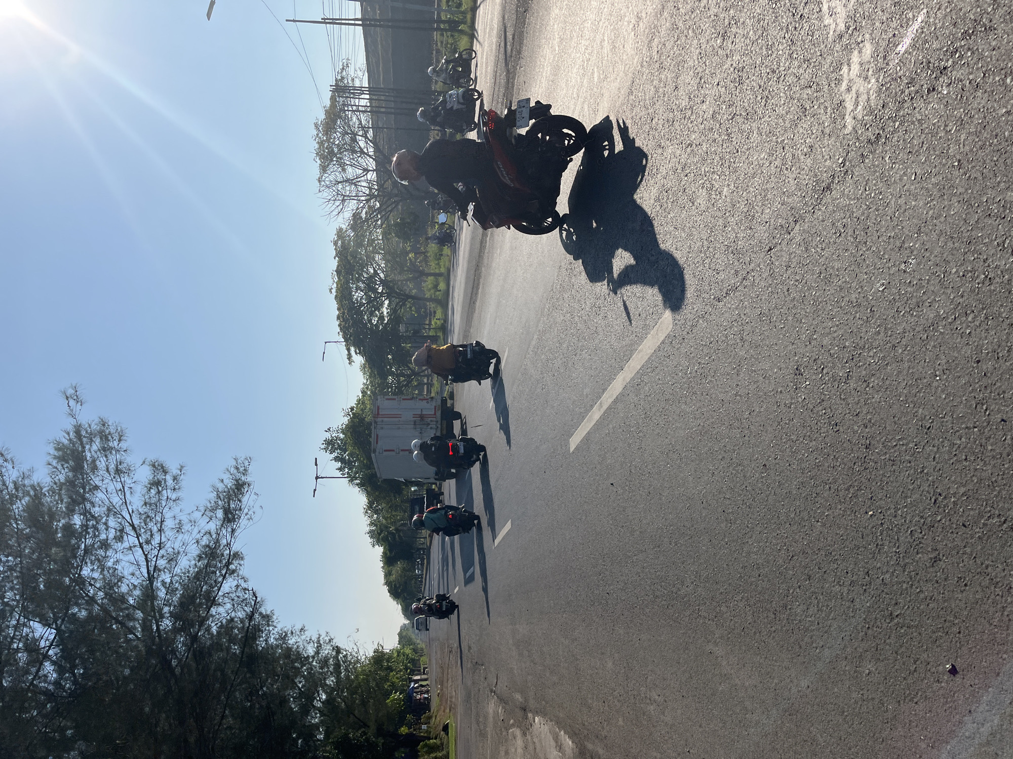 The image depicts a road in the city of Siring, Indonesia, bathed in the warm glow of a late afternoon sun. Several motorcycles, each with one or two riders wearing helmets, are traveling on the asphalt road. The riders appear to be heading in the same direction, indicated by the composition of the image. Trees line the left side of the road, casting shadows, and in the distance, a white truck with a container is visible. The sky is clear and blue with the sun creating lens flare effects. The overall scene conveys a sense of everyday life and transportation in a tropical urban setting.