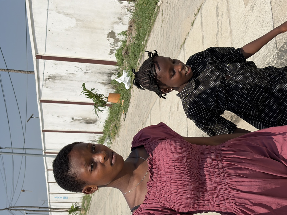 The image displays two females walking outdoors in Jakande, Nigeria, on a sunny day. In the foreground, a young woman with dark skin and short black hair wears a dark red, off-the-shoulder dress with a shirred bodice and a thin silver necklace. She faces forward, looking directly at the viewer with a neutral expression. To her right and slightly behind her, a younger female child with dark skin and braided hair styled in multiple small upright sections is visible. This child wears a black collared shirt with white polka dots and looks downwards and to her right, displaying a slight open-mouthed smile. They are walking on a light-colored, paved pathway. The background consists of a tall, weathered white concrete wall featuring vertical dark brown segments. A narrow strip of green grass with sparse plants, including one in an orange pot, runs along the base of the wall. A crumpled white object, consistent with litter, rests on the grass. Overhead, thin utility poles and wires are visible against a clear, light blue sky.