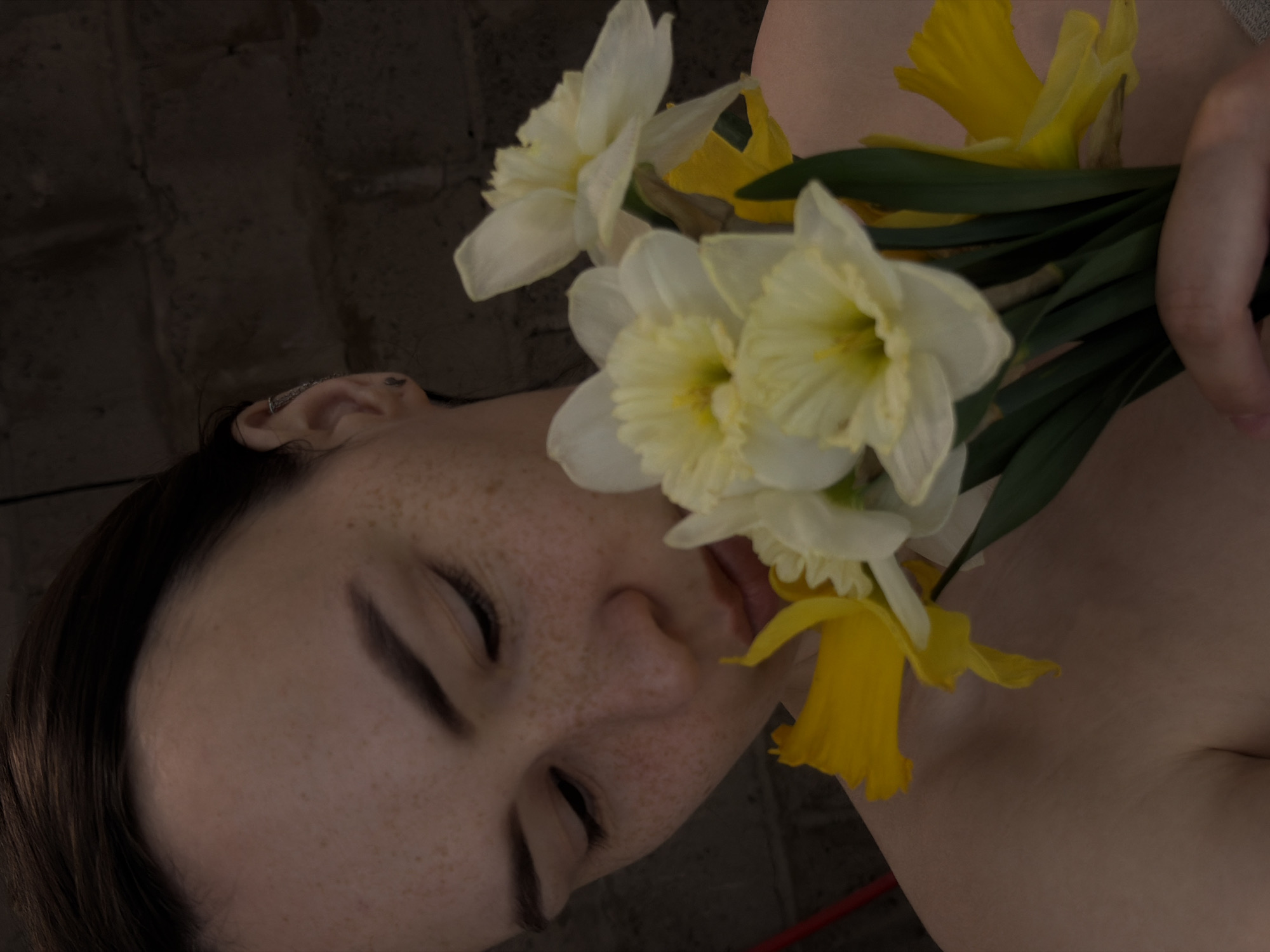 A close-up shot features a person with closed eyes and freckled skin, holding a bouquet of white and yellow daffodils. The person's face is angled, with their hair visible on the left side. An earring is present in their ear. The bouquet is positioned near their face, with some of the flowers partially covering their mouth. The background is a dark, textured surface, possibly a wall. A portion of bare skin is also visible. There is no information in the image to confirm it was taken in Chaikovsky, Russia.