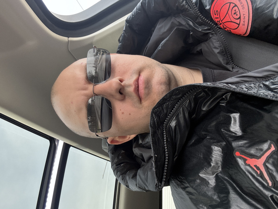 The image features an adult male seated inside a vehicle, located in Yasen, Bulgaria. The subject is bald, has fair skin, and looks directly at the camera. He wears rimless, rectangular sunglasses with dark lenses, reflecting an exterior scene of a road lined with trees under a bright or overcast sky. He is dressed in a black, shiny, puffy jacket with a visible zipper along the collar. A red "Jumpman" logo is present on the left side of the jacket, and a circular red "PARIS SAINT-GERMAIN" patch is on the right. The vehicle's interior shows a light-colored headliner and an open sunroof with a white illuminated strip positioned above the subject's head. No other individuals or specific landmarks identifying Yasen are discernible within the frame.