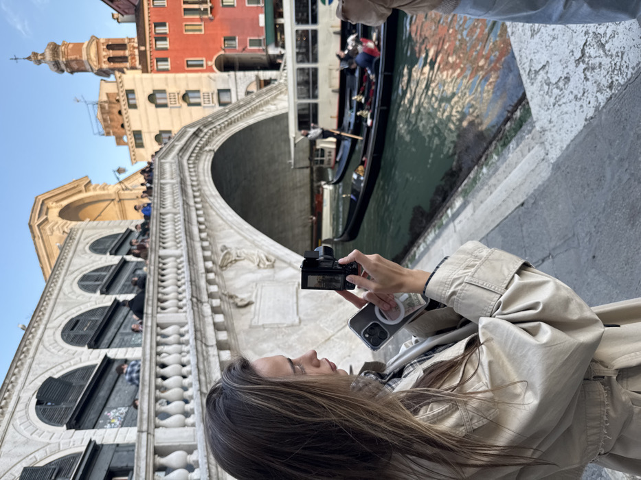 The image depicts a tourist scene at the Rialto Bridge over the Grand Canal in Venice, Italy. In the foreground, a young woman with long brown hair, wearing a beige trench coat over a plaid shirt, is seen in profile from the right. She is holding a black compact digital camera in her right hand, pointing it forward, and her left hand holds a clear smartphone with a white circular attachment. Her gaze is directed towards the camera screen, indicating she is actively taking a photo or video.
Behind her, the stone Rialto Bridge spans the Grand Canal, featuring multiple arches and a balustrade along its upper deck where numerous people are visible. Shops with dark shutters are integrated into the bridge's structure. On the Grand Canal, directly under and beyond the bridge, a dark gondola with at least two passengers and a standing gondolier is present, propelled by an oar. A white public vaporetto is also visible further down the canal. In the background, a cluster of multi-colored buildings, including yellow and red structures, rise above the canal, topped by a distinct ornate bell tower featuring an arched opening and a cross at its apex. The sky is clear blue with soft daylight.