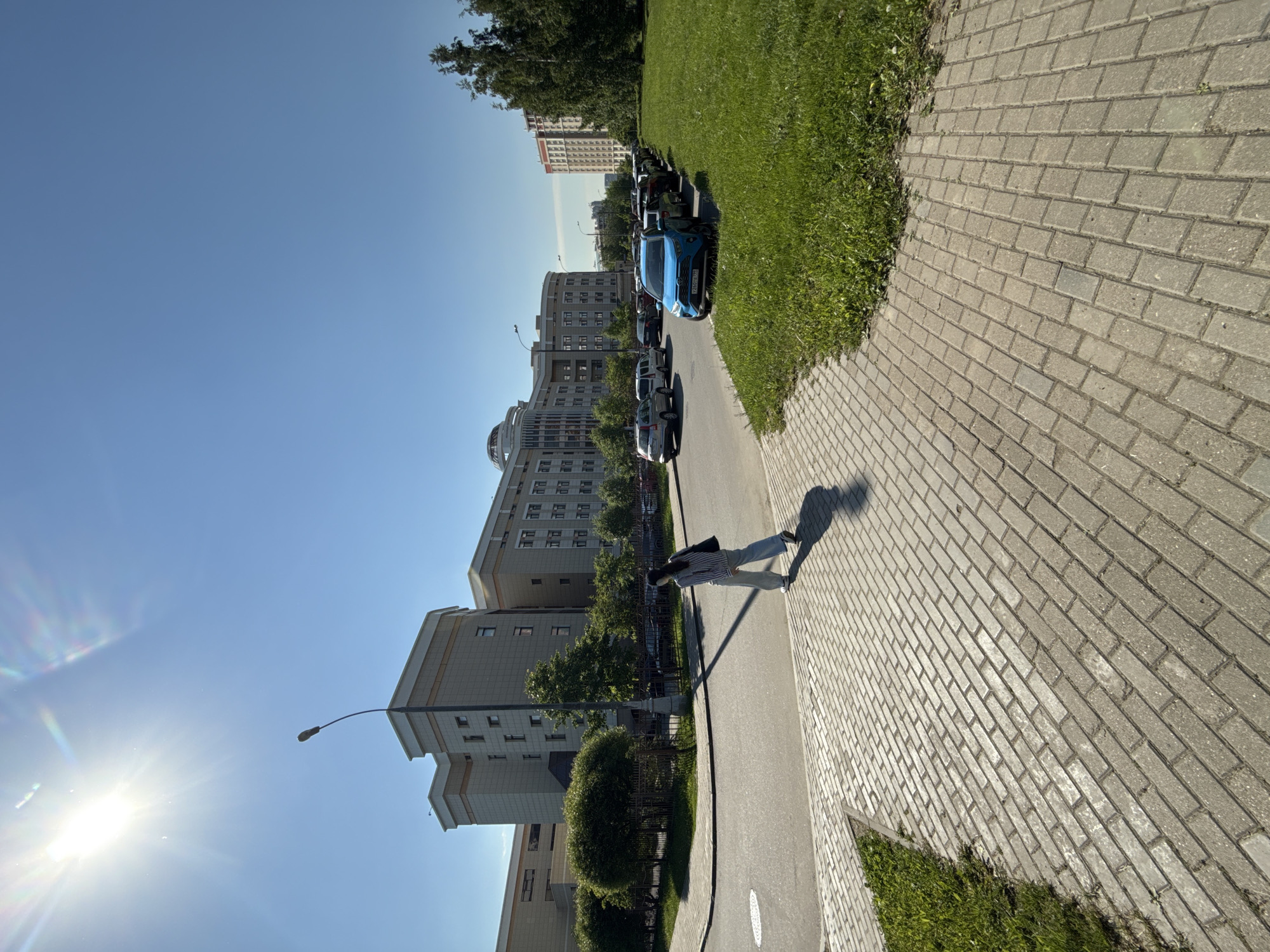 This image captures a vibrant, sunlit urban street scene in Moscow, Russia, oriented as if looking down a sidewalk. A broad paved sidewalk occupies the right foreground and middle ground, bordered by a strip of lush green grass. Further to the left, an asphalt road is lined with numerous parked cars. Beyond the road, several multi-story, modern-looking buildings rise, interspersed with trees and manicured green spaces.
A lone individual, appearing to be a woman, walks away from the viewer on the paved sidewalk. She is dressed in light-colored clothing and casts a noticeably long shadow stretching out in front of her, indicating the sun is low in the sky, likely early morning or late afternoon.
Among the parked vehicles, a distinctive blue SUV or crossover stands out in the mid-ground. Its Russian license plate is clearly visible, reading "X931AA 799 RUS," with the "799 RUS" code confirming the location as Moscow. The buildings lining the street feature contemporary architecture, suggesting a residential or commercial district. A lamppost is also visible extending from the buildings towards the sky.
The weather is bright and clear, with an expansive, cloudless blue sky dominating the upper portions of the frame. The sun, positioned in the lower-left, causes a prominent lens flare, adding a radiant quality to the image. The scene depicts a routine daily moment, with no specific event or overt emotions expressed.