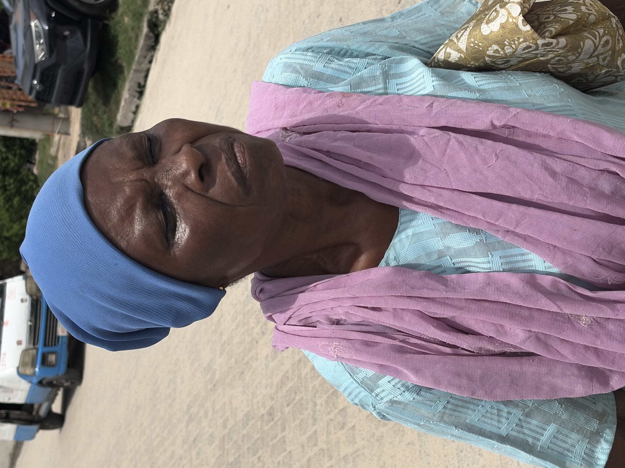 A close-up shot depicts an elderly Black woman wearing a blue head covering. A light blue blouse with a geometric pattern is visible underneath a loosely draped, light purple scarf. A patterned, gold-toned pillow is positioned behind her head. The woman's eyes are closed, and her face appears relaxed. The setting appears to be outdoors in Igboefon, Nigeria. A vehicle and truck can be seen in the background, slightly out of focus. The ground is light-colored, possibly concrete or pavement.