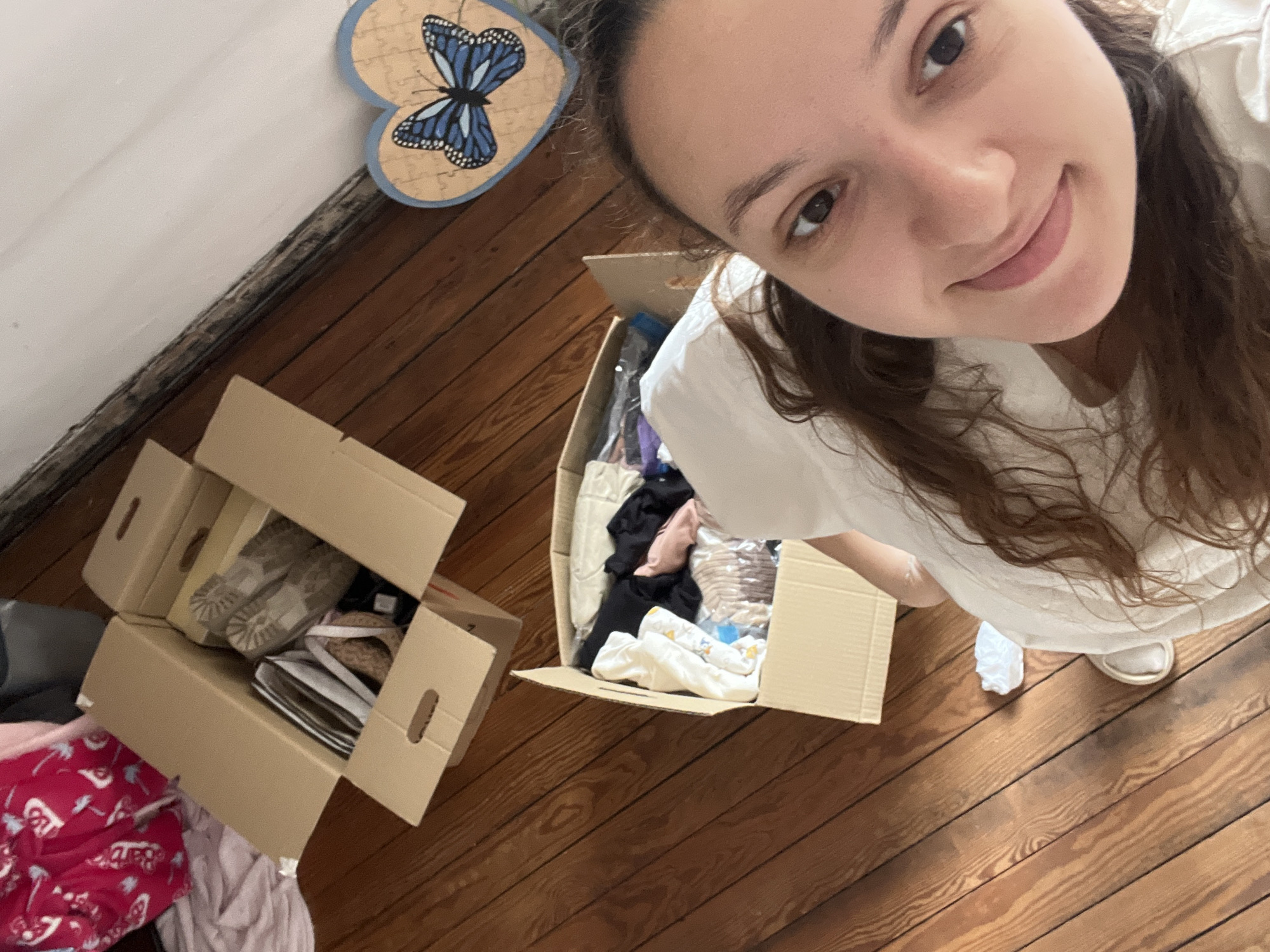 A selfie shows a young woman standing above two cardboard boxes. One box contains shoes, and the other contains clothes and plastic bags. The location appears to be indoors, evidenced by a wooden floor, white wall, and a wooden butterfly decoration on the wall. The environment suggests a residence, possibly during packing or unpacking. No city information provided.