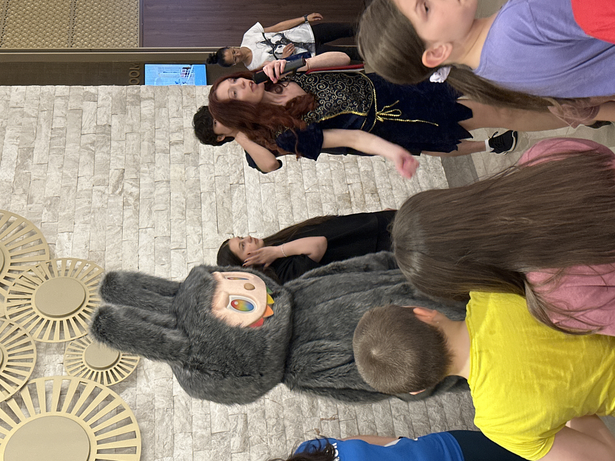 The image shows a group of people gathered in what appears to be a public space, possibly a lobby or event hall. A woman with auburn hair is standing and appears to be speaking into a microphone. Several children and adults are present. A large, gray, furry mascot-like figure with an unusual face is lying on the ground near the group. The background consists of a light-colored, textured wall and decorative golden circular shapes. In the upper left of the image, a sign with the word "ROOM" is visible. The location is Kazanlak, Bulgaria.