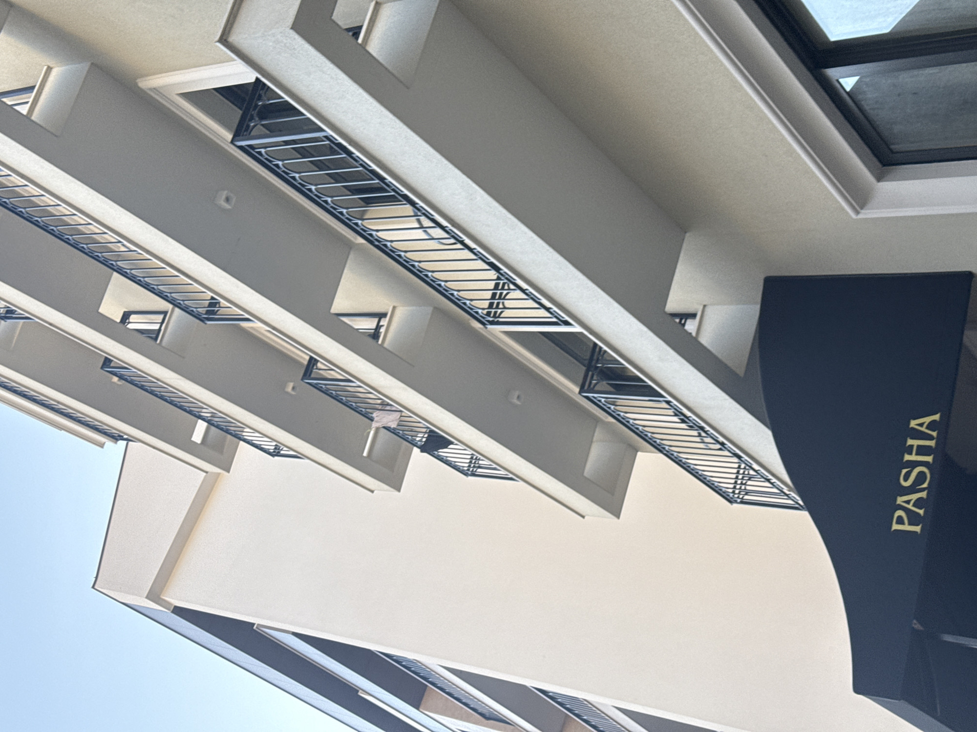 The image displays the exterior facade of a multi-story building in Budva, Montenegro, captured from a low-angle perspective. The building is characterized by a cream-colored stucco finish and features multiple cantilevered balconies stacked vertically. Each balcony is equipped with a black, ornamental wrought-iron railing consisting of arched vertical balusters. On the second visible balcony from the top, on the right side, light-colored fabric is draped over the railing. At the base of the building, a dark, possibly black, fabric awning is visible, emblazoned with the word "PASHA" in gold capital letters. A section of an adjacent or connected building, with a darker, possibly gray, upper facade and similar balconies, is partially visible on the left. The sky is clear and light blue, indicating daylight conditions. No individuals or active interactions are present in the scene.