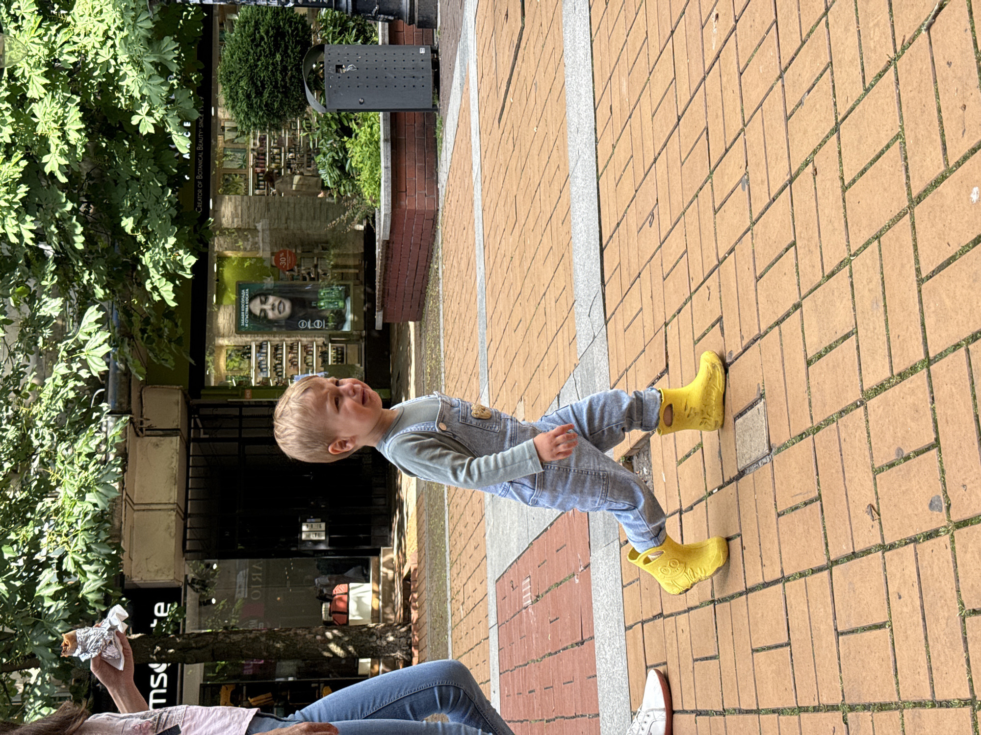 This image depicts an outdoor urban scene, likely a pedestrian street or shopping area, on a bright, sunny day in Sofia, Bulgaria. The pavement consists of light brown rectangular brick pavers, with some grass or weeds growing in the interstices, and a strip of lighter grey stone running through it.
The primary subject is a young child, appearing to be a toddler, with blond hair, dressed in a light green long-sleeved shirt and light blue denim overalls. The child is wearing distinctive bright yellow rain boots or clogs that feature a design resembling an animal's face. The child's mouth is open, suggesting they are in the midst of crying or expressing a strong emotion.
In the bottom-left corner, part of an adult's body is visible, seated. This person is wearing blue jeans and white and red sneakers, and holds an item wrapped in aluminum foil, possibly food. Their legs are positioned near the child's feet.
The background features several storefronts with large glass windows, reflecting elements of the street and sky. One prominent store sign is partially visible, showing "CREATOR OF BOTANICAL BEAUTY SINCE 1959" (identifiable as Yves Rocher). Another advertisement within a storefront displays a woman's face, Bulgarian text "ЗАЕДНО КУПУВАМ ЗА И СЪСТАВКИ КОГАТО" (translating roughly to "Buying together for ingredients when..."), and a "-30%" discount sign. Green foliage from trees and bushes lines the left side of the street and appears in reflections, contributing to the daytime ambiance. A black trash can is also visible in the background.