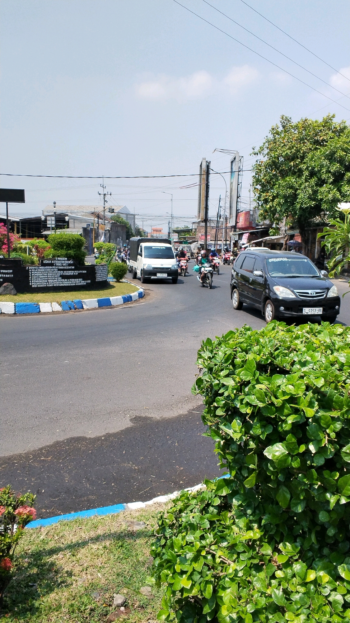 The image depicts a street scene in Banjaragung, Indonesia, on a bright, sunny day. A multi-lane road curves to the left, with vehicles navigating the intersection. In the foreground, lush green bushes flank the road and a grassy median. A white truck is in motion on the road, followed by several motorcycles carrying riders. To the right, a black minivan also travels along the road. The background reveals a mix of commercial buildings, power lines, and mature trees, indicating an urban or semi-urban environment. A signpost with text is visible on the left side of the road, and a traffic island is adorned with blue and white painted curbs and manicured plants. The sky is clear with a few scattered clouds.