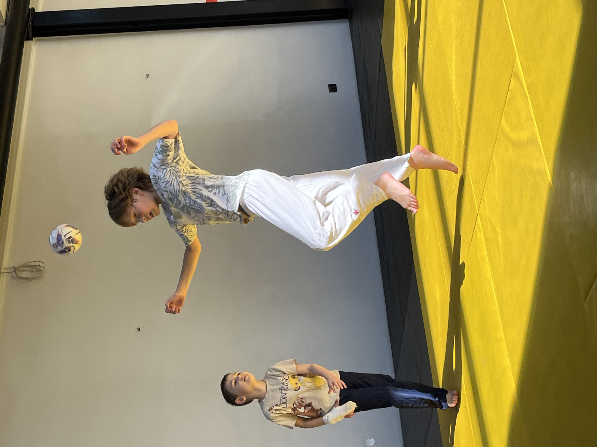 A scene indoors, likely a gym or recreational space, features a person suspended horizontally in mid-air, dressed in a white top with a leaf print and white pants, with bare feet. The person appears to be in motion, possibly kicking a soccer ball positioned to the left. A second person, a child wearing a "Lion King" T-shirt and dark pants, lies on the floor looking up towards the suspended individual. The environment consists of a light-colored wall and a yellow, textured surface, possibly mats or padding, against which sunlight casts shadows. The location, Krasnoyarsk, Russia, is not evident from the visual details.