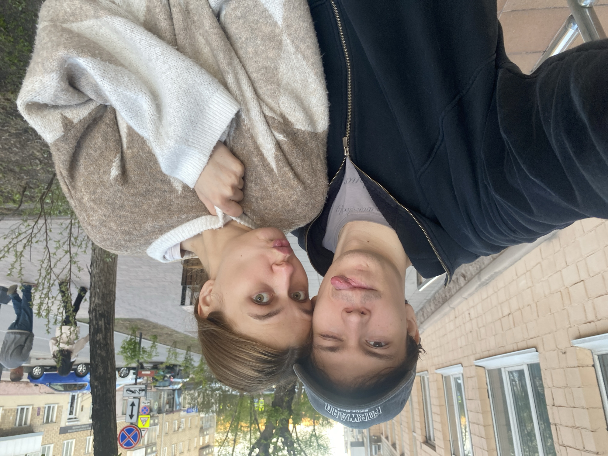 A selfie taken by a male and female near the intersection of Mira Avenue and Surikova Street in Krasnoyarsk, Russia. The female is on the left, wearing a tan and white sweater and pursing her lips. The male is on the right, wearing a black zip-up hoodie and a dark cap with the word "FORWARD" visible. He is sticking his tongue out and winking. The background shows a city street with buildings, trees, road signs and parked cars. There are people walking on the sidewalk. The image is rotated 180 degrees.