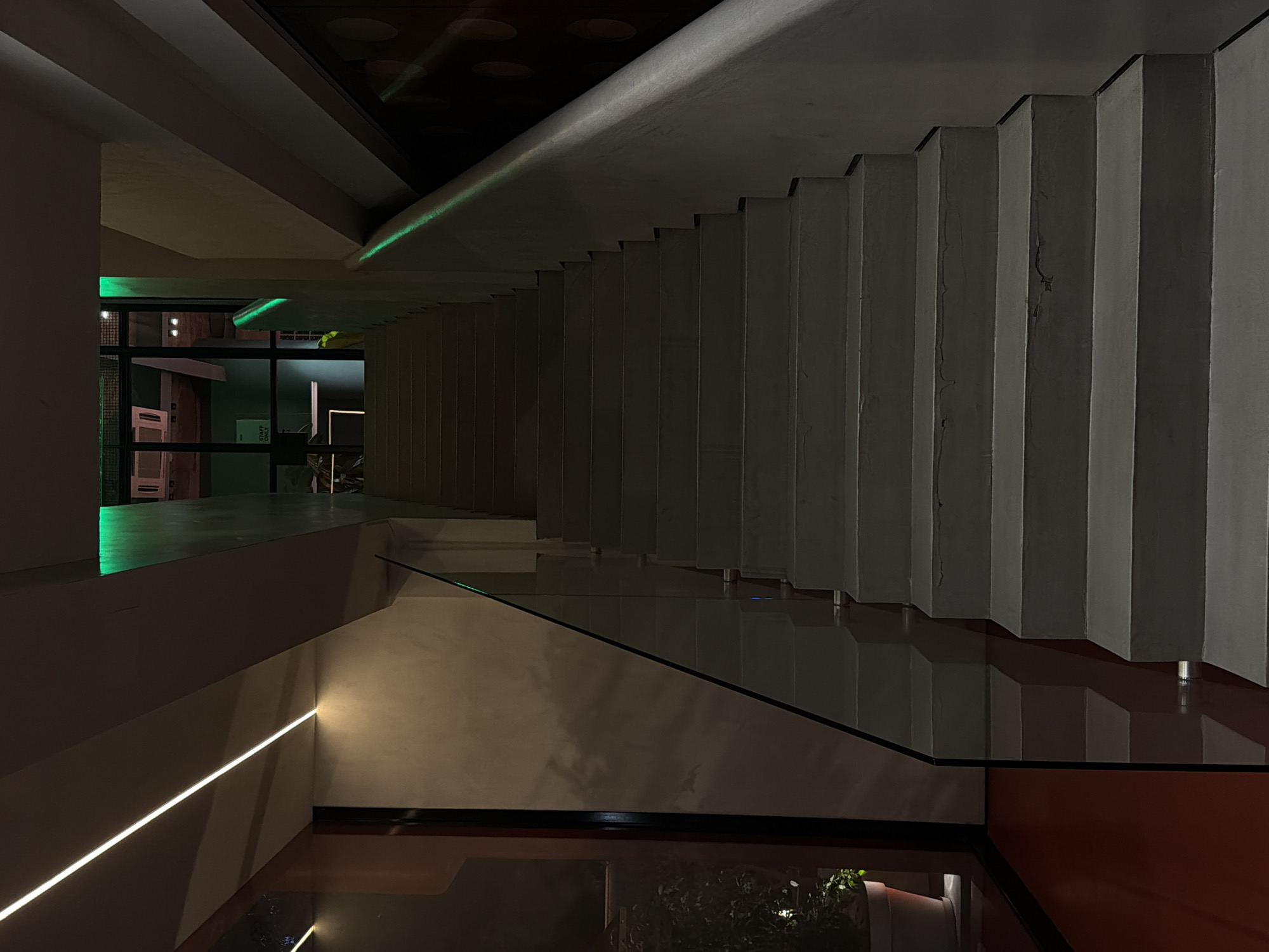 The image displays a modern, minimalist interior architectural space, featuring a prominent staircase and striking artificial lighting. The scene appears to be set at night or in a dimly lit environment, highlighting the interplay of light and shadow.
The central subject is a robust, geometric staircase made of concrete or a similar material, with a distinctive stepped underside that recedes diagonally from the upper right toward the center. A sleek glass railing runs along the edge of an upper landing and descends alongside the staircase, adding to the contemporary aesthetic.
Illumination is provided by LED strip lighting. A vibrant green LED strip is visible along the underside of the upper landing and possibly portions of the staircase, casting a futuristic glow and reflecting on the glass surfaces. A warm white LED strip illuminates a lower wall on the left, creating a clean, defined line of light.
The glass railing and potentially a dark, reflective floor below create reflections of the staircase's steps and the green light, adding depth to the composition. The dominant color palette consists of cool grays from the concrete, contrasted by the vivid green and soft white artificial lights. In the lower right foreground, a hint of dark reddish-brown adds a subtle warmth.
Through a window or glass panel in the background on the left, a sign is faintly visible with the text "STAFF ONLY." There are no people present in the image, emphasizing the architectural design and the controlled lighting environment.