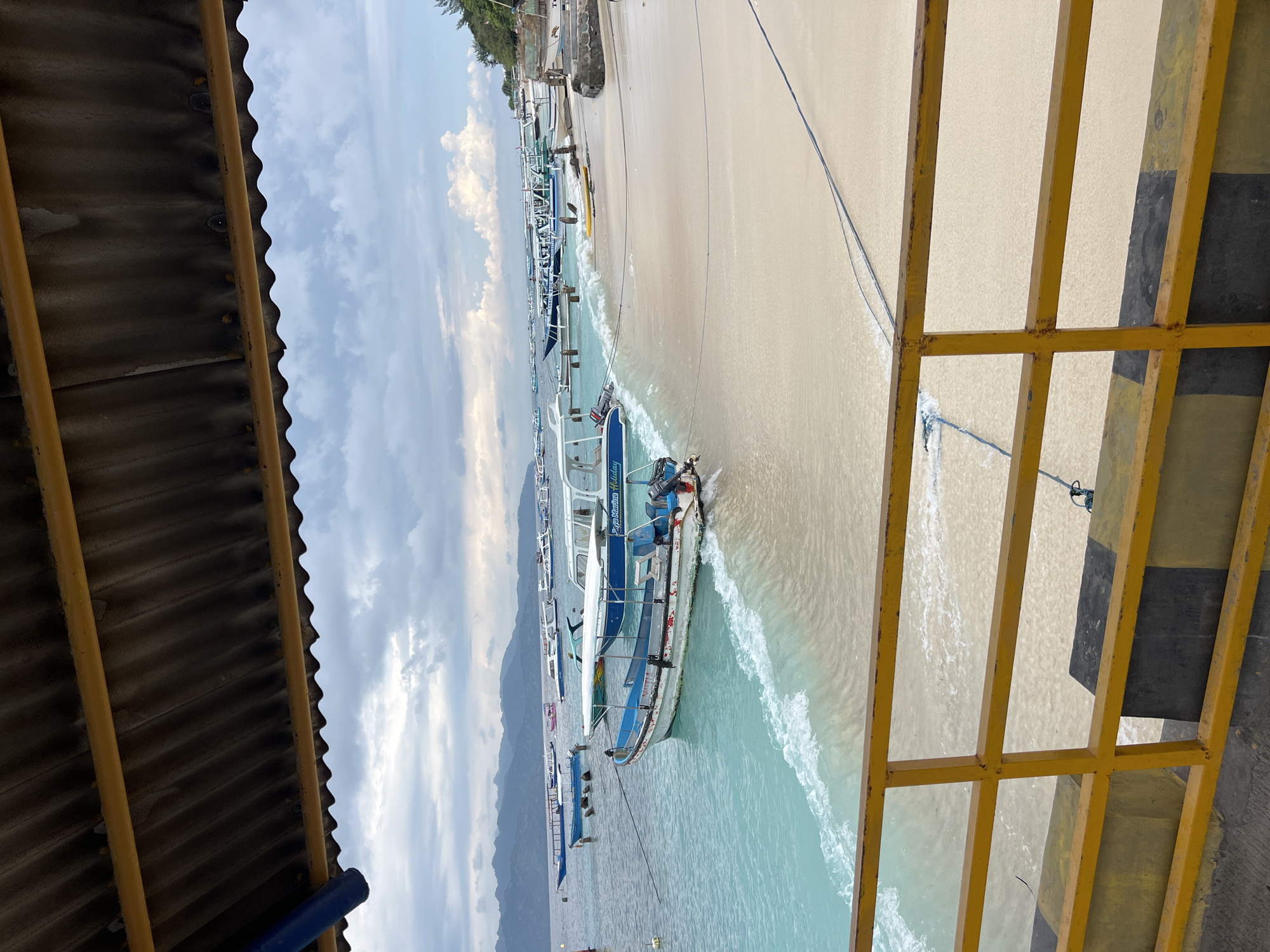 A horizontal, medium-shot image captures a beach scene in an undisclosed location in Indonesia. The scene is framed on either side by an awning on the left and a yellow metal structure on the right, suggesting the perspective is from a building or structure overlooking the beach.
The beach is populated with several boats anchored in the shallow water near the shore. The water is light blue, transitioning to white where the waves are breaking. The sand is light beige.
The sky is overcast with clouds, and mountains are visible in the distance. The overcast sky suggests a cloudy day. No people are visible in the frame.