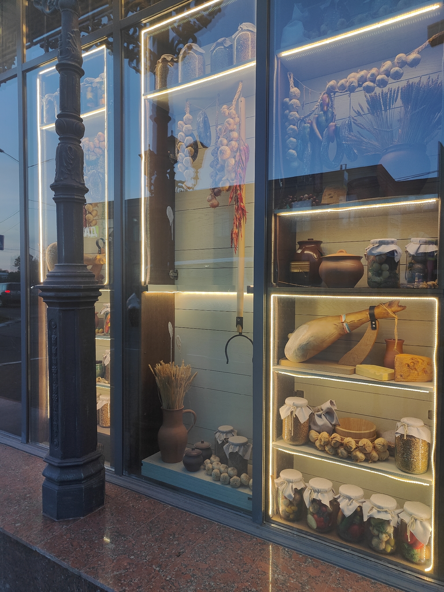 This is a medium shot, eye-level, outdoor, daylight photograph of a store window display. The display features various preserved food items and cured meats. On the left, a tall, dark, ornate lamppost stands in front of the window. The window itself is divided into several illuminated shelves. The top shelf displays jars of what appear to be grains or seeds. The shelf below it holds two dark ceramic pots and several jars of olives and other pickled items. The next shelf down features a large cured ham leg mounted on a wooden stand, accompanied by a small pitcher and a wedge of cheese. The bottom shelf is filled with jars of pickled vegetables. Dried garlic bulbs and peppers hang from strings in the background, along with bundles of wheat. The store's facade is constructed of light-colored wooden planks. The ground in front of the store is a speckled red granite. The overall impression is of a rustic, artisanal food shop.