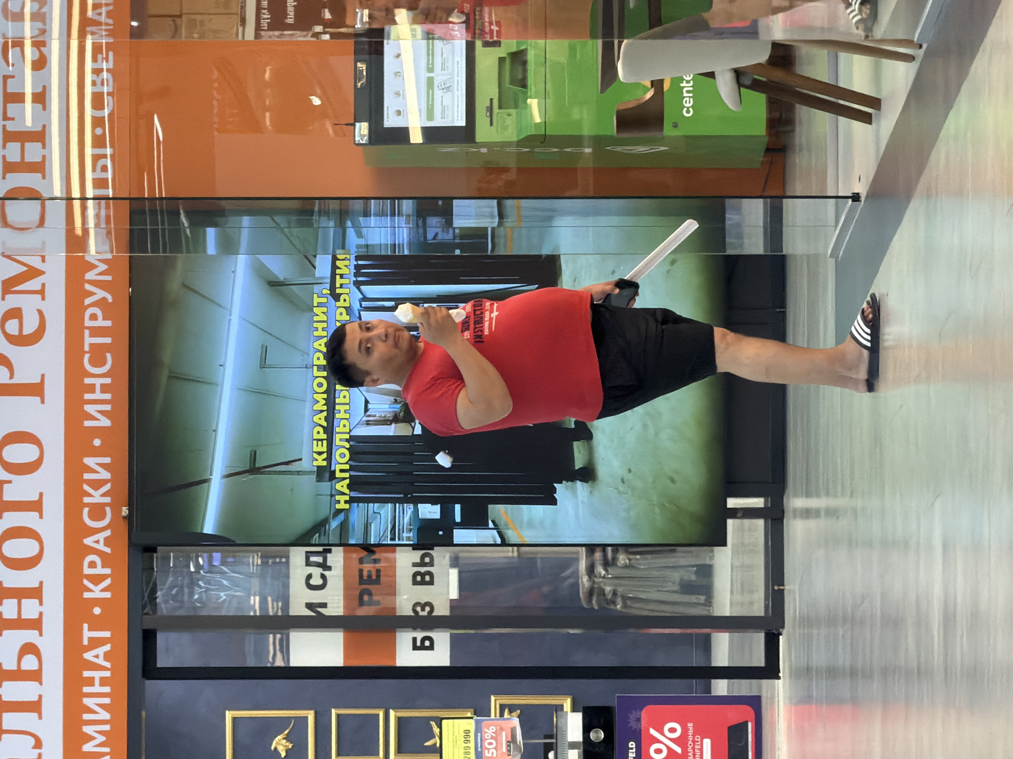 The image captures a street scene, possibly in Astana, Kazakhstan, though the location is not explicitly confirmed. A man is the central subject, wearing a red t-shirt and black shorts, with Adidas slides on his feet. He is holding an ice cream cone in his right hand and a white object, possibly a ruler or a long, flat tool, in his left hand. He stands near a glass partition, and a large digital screen is visible behind him, displaying text in Russian that appears to be advertising. Other elements in the background include what looks like retail displays, a green kiosk or machine, and furniture. The overall environment suggests a commercial or public space, perhaps a shopping center or a hardware store, with a glossy floor reflecting the ambient light.