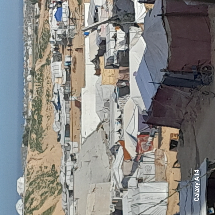 A crowded refugee camp spans across a sandy landscape, possibly a coastal area. Makeshift tents and shelters dominate the scene, constructed from various materials. Clotheslines with hanging laundry are visible between dwellings. The sea is visible in the distance. Overall impression suggests challenging living conditions due to overcrowding.