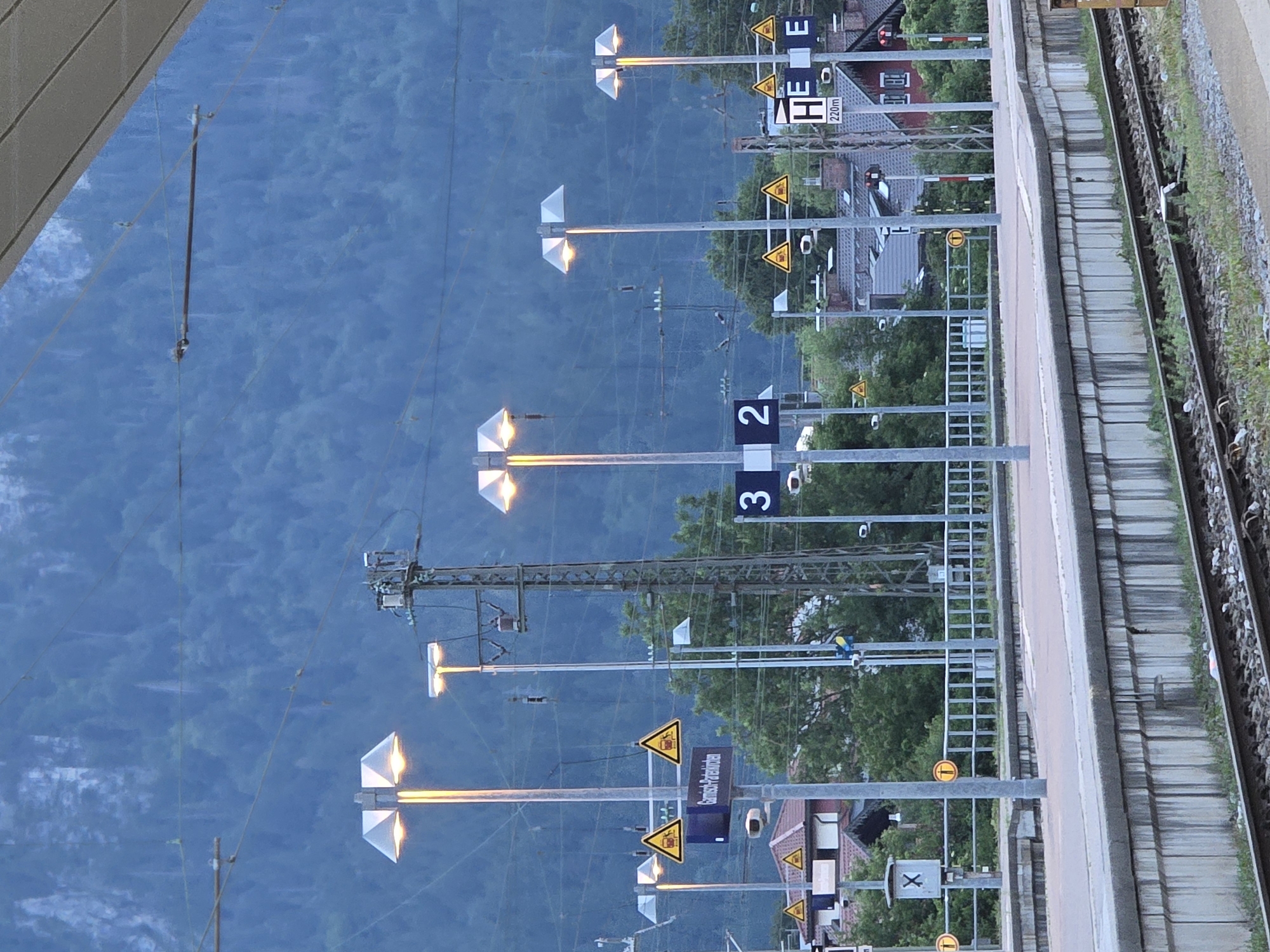 The image shows a train station platform in Garmisch-Partenkirchen, Germany. The platform is made of concrete and has a metal railing. There are several lampposts with modern, angular shades casting a warm light. In the background, there is a dense forest covering a steep mountain slope, characteristic of the Bavarian Alps. Several signs are visible, including triangular warning signs with a symbol of a train, and a blue sign with "Garmisch-Partenkirchen" written on it. There are also platform number signs displaying "3" and "2". Overhead wires for the train power system are visible, along with electrical infrastructure like transformers and poles. The overall scene is serene, with the warm lighting of the lamps contrasting with the cool, muted colors of the natural landscape and the twilight sky. No people are visible in the image.
