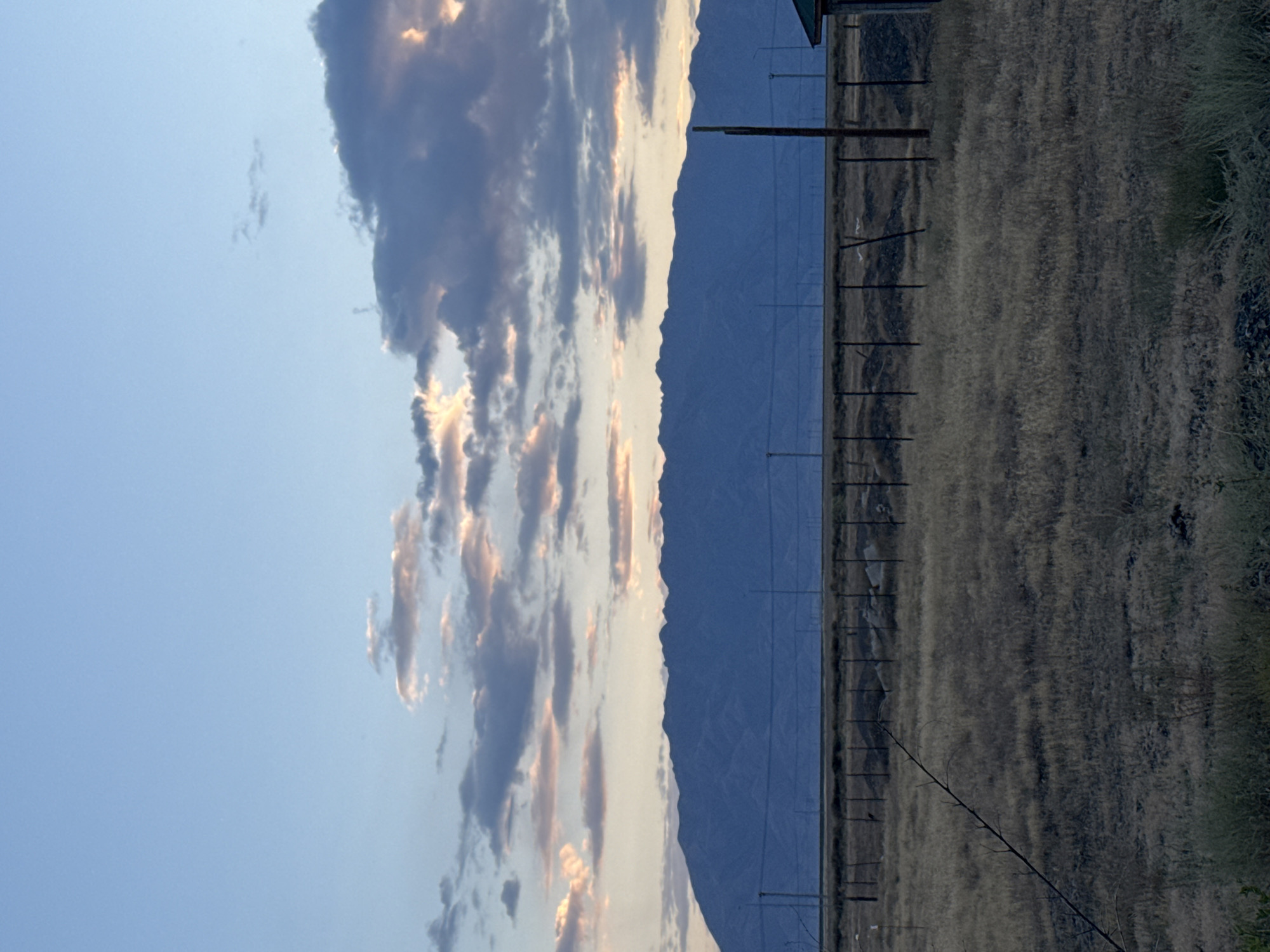 A wide shot captures a vast, arid landscape with a prominent mountain range in the background under a hazy, twilight sky. The sky displays a gradient of blues and purples, with streaks of orange and pink illuminating scattered clouds. A dark, jagged mountain range stretches across the horizon, its peaks softened by distance and atmospheric haze. In the mid-ground, a chain-link fence runs horizontally, separating the foreground from a field of dry, yellowed grass. Several dark, vertical fence posts are visible, some topped with lights. To the far right, a corner of a dark green, angular building is partially visible. The foreground consists of more dry grass, with a thin, dark, leafless tree branch extending diagonally from the lower left corner. The overall impression is one of a remote, undeveloped area experiencing the transition from day to night.