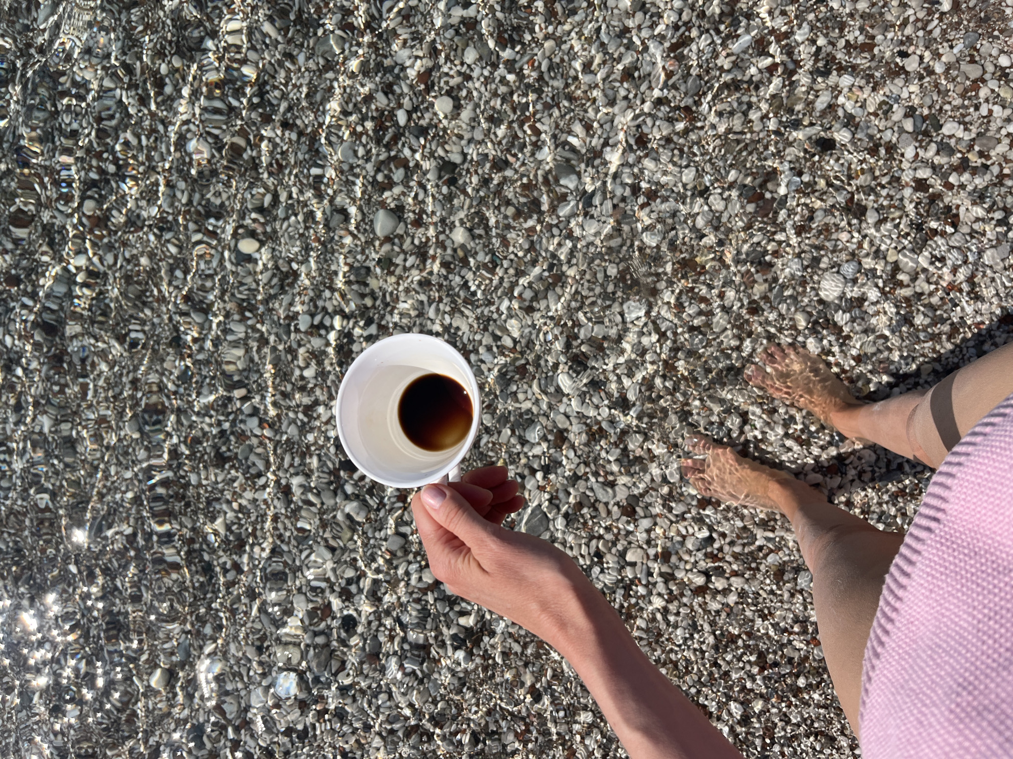 A high-angle, sunlit shot shows a woman holding a white cup of dark liquid (likely coffee) over clear, shallow water on a pebble beach. The water is clear enough to see the stones beneath. The woman's feet are partially submerged, and a portion of her pink top is visible on the right side. The location is possibly a beach in or near Budva, Montenegro, judging by the typical Adriatic coast features of pebble beaches and clear water.