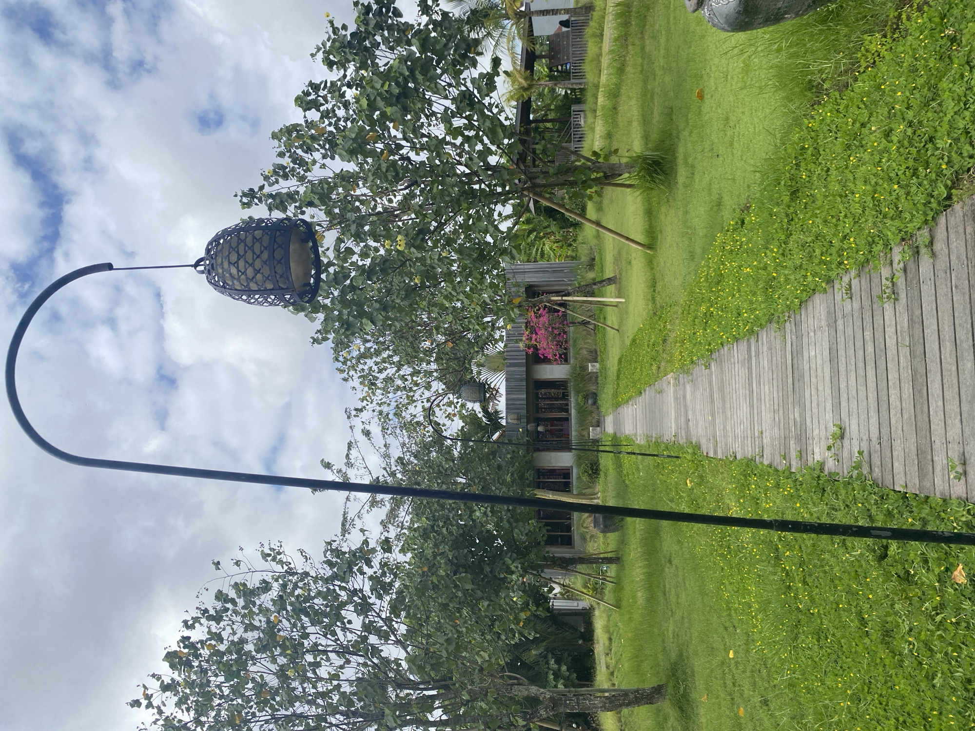 A wooden plank walkway stretches into the distance, flanked by green grass and low-lying vegetation with small yellow flowers, in North Kuta, Indonesia. A black, shepherd's crook-style metal lamppost with a woven cage lantern hangs suspended over the walkway. The sky is overcast with white clouds. Trees are on either side of the walkway, partially obscuring a building in the background. The image is oriented sideways.