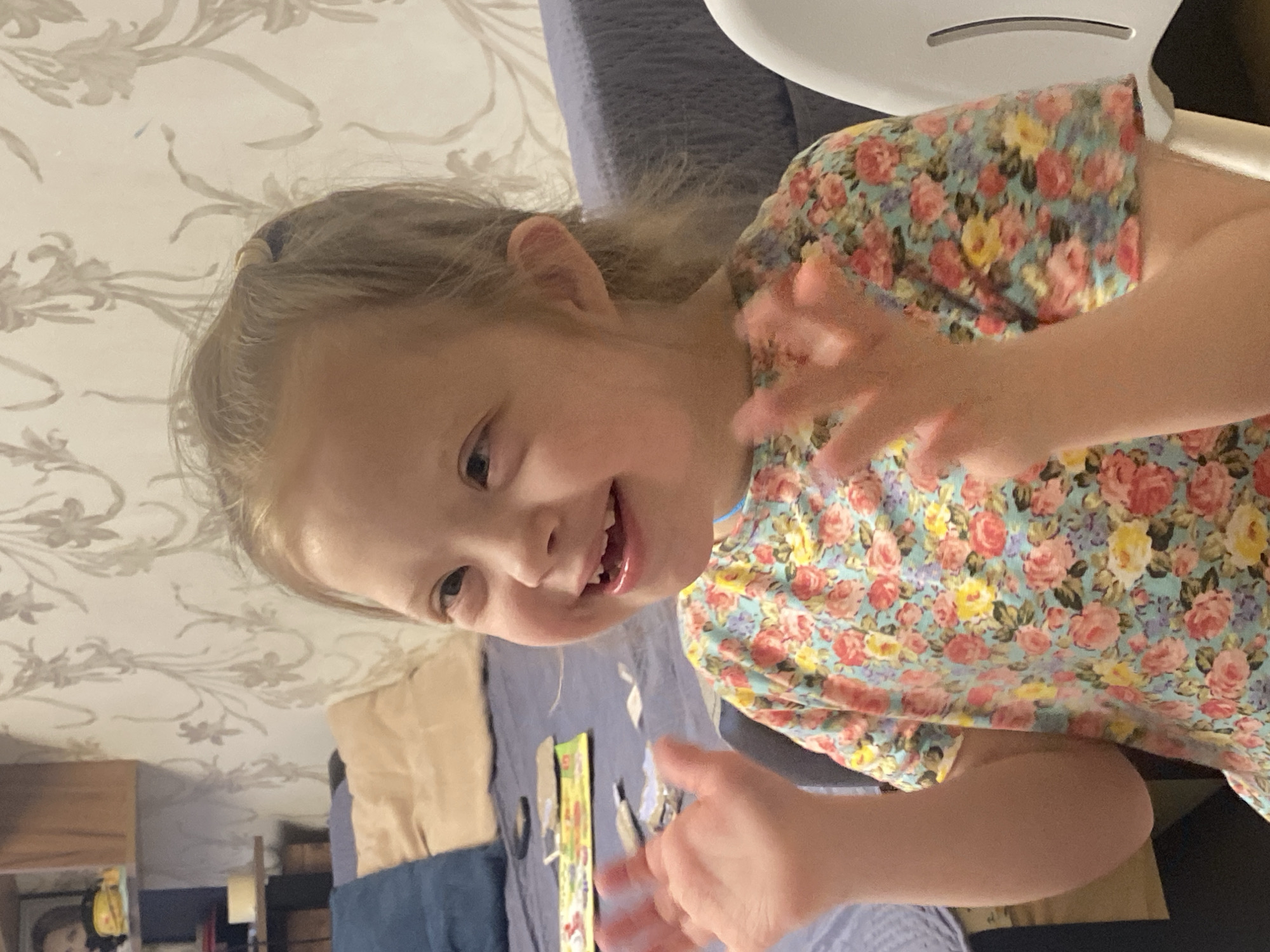 A young girl with light hair is smiling and gesturing with her hands toward the camera. She is wearing a floral-patterned dress. The background shows a room with a patterned wallpaper, furniture, and what appears to be craft supplies on a surface. There is no evidence to suggest this image was taken in Yekaterinburg, Russia.