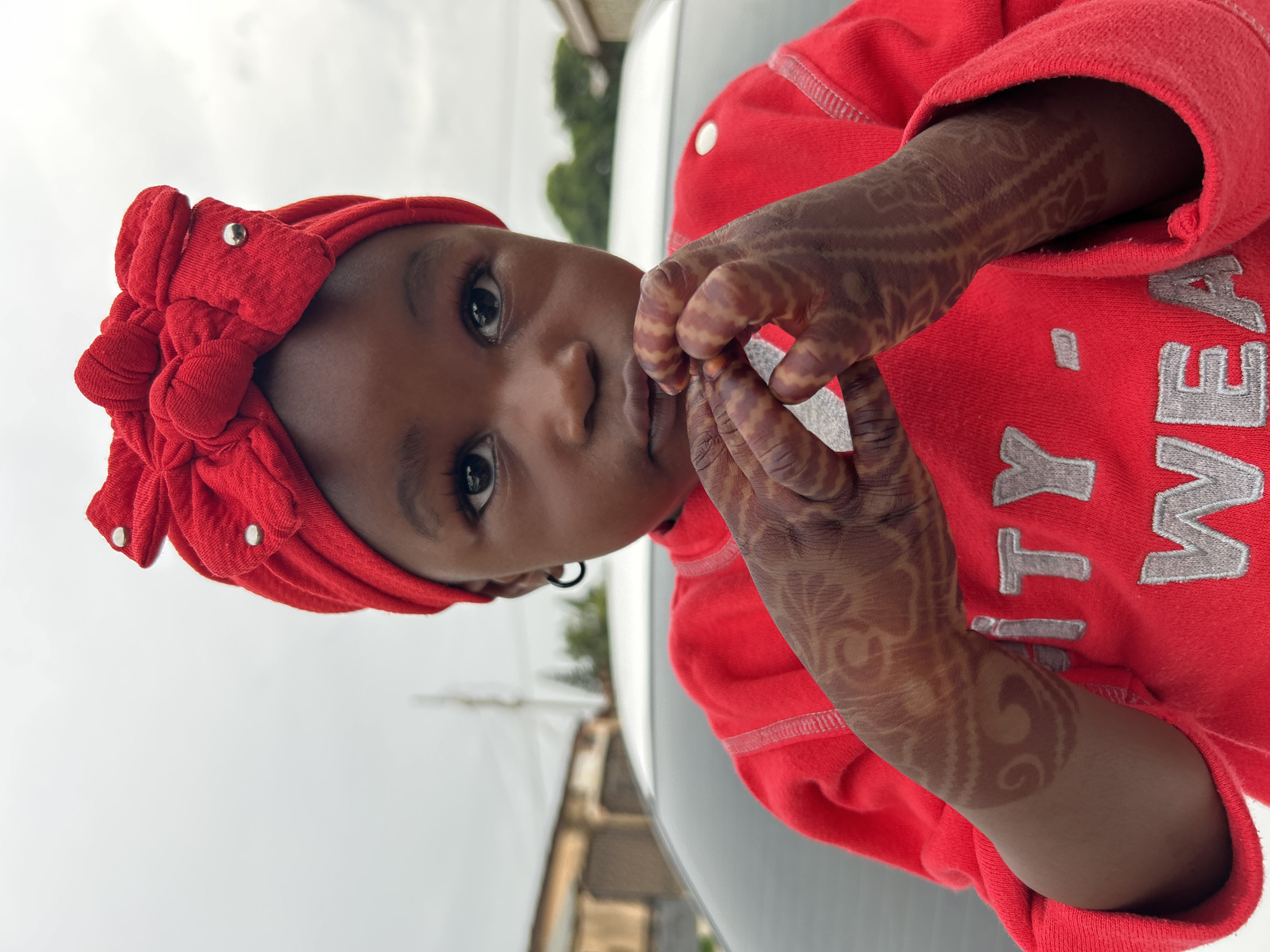 A young child with brown skin is pictured close-up, likely in Jos, Nigeria. The child wears a red knotted head wrap and a red long-sleeved shirt with silver lettering. Intricate henna designs adorn the child's hands and arms. The child is looking towards the camera with a neutral expression. The background shows a slightly overcast sky and blurred buildings and foliage, suggestive of an urban environment. A white car is also partially visible.