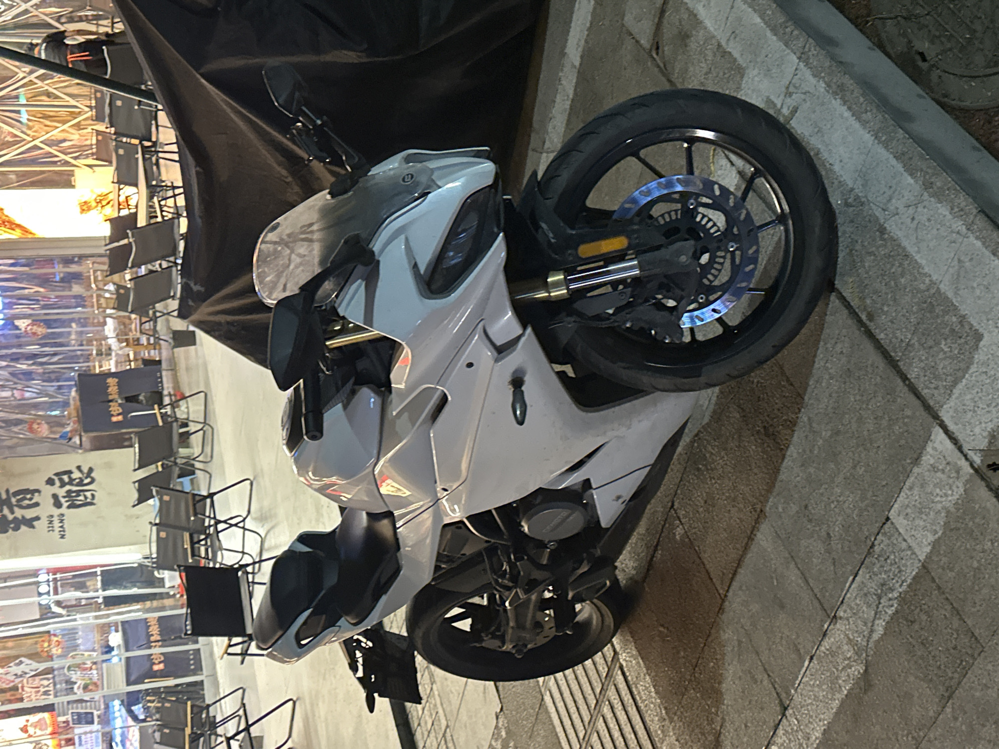 A white sport motorcycle is parked on a paved sidewalk or pedestrian area, leaning on its kickstand. The motorcycle is the central subject, positioned diagonally across the frame, showcasing its sleek design, front fairing, handlebars, and front wheel. The front brake disc exhibits a noticeable blueish, iridescent sheen.
The setting appears to be an urban environment at night or dusk, illuminated by artificial lights emanating from the background. To the left of the motorcycle, a row of black folding chairs and tables, possibly belonging to a street-side restaurant or food stall, are visible. Above these, a large black tarp or tent structure covers a portion of the area. Further in the background, bright lights and reflective surfaces suggest commercial establishments or buildings.
Visible text on signs and banners in the background includes Chinese characters that transliterate to "JING NIANG," and other characters like "烤肉" (kǎo ròu, meaning barbecue) and "江湖" (jiāng hú). These textual cues, along with the overall urban aesthetic, firmly place the scene in China, aligning with the provided context of Chongqing High-tech Zone. The motorcycle appears to be in good condition, blending into the lively night-time street scene.