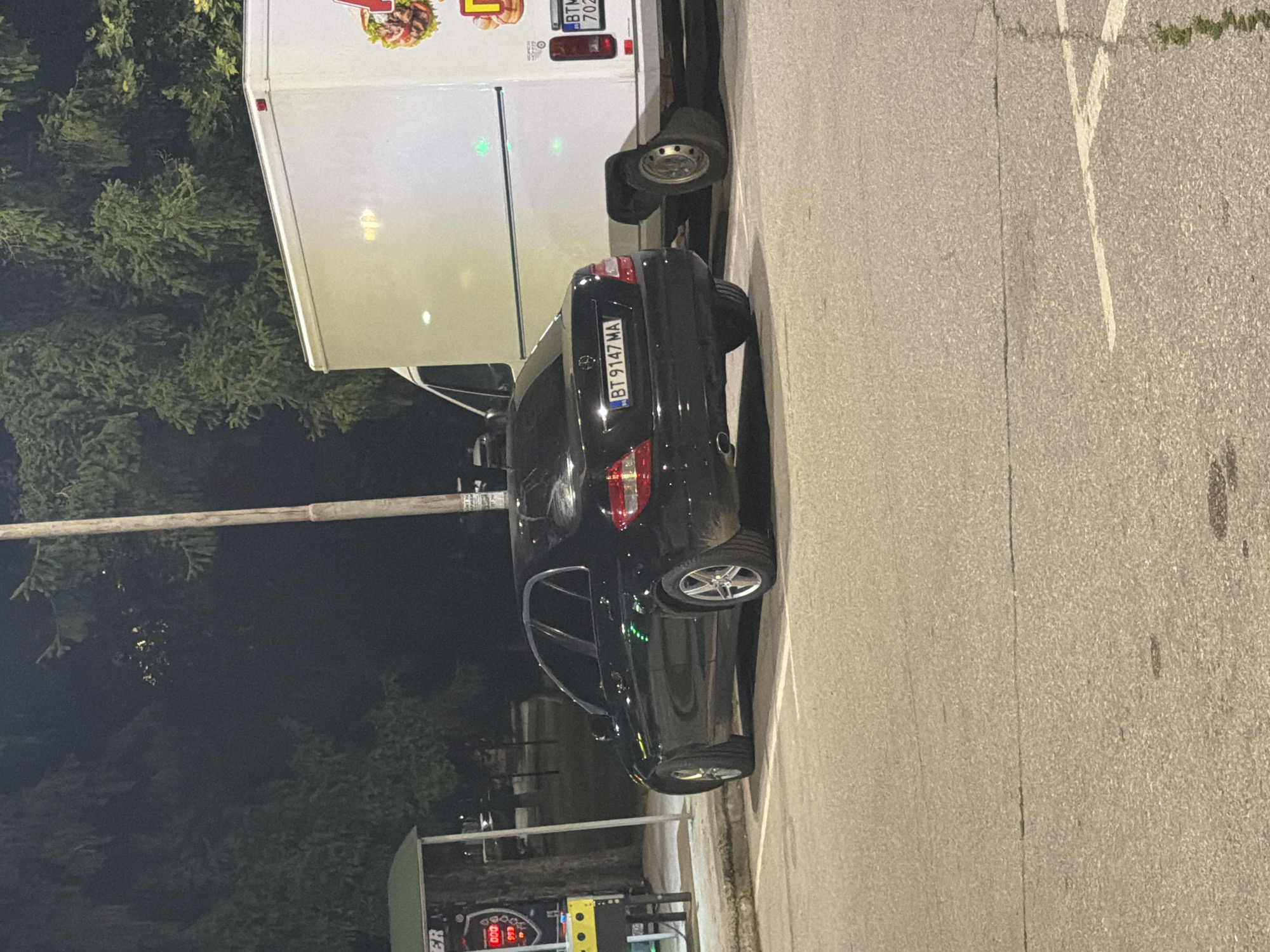 The image shows a black Mercedes-Benz C-Class sedan parked in a lot at night, with a white food truck behind it. The car's license plate reads "BT 9147 MA". To the left of the car is a gas pump. The scene appears to be a public parking area, possibly near a roadside establishment given the presence of the food truck. The background consists of trees and a lamppost. The location is identified as Strazhitsa, Bulgaria.