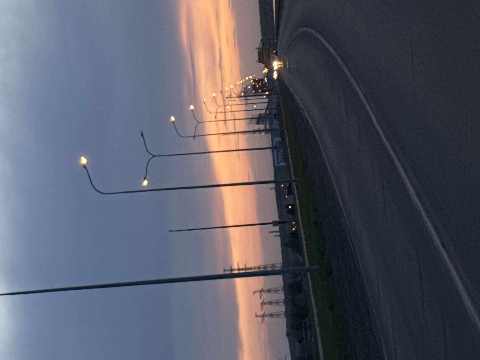 The image shows a view of a road and a row of street lights at twilight in Chaikovsky, Russia. The sky is colored with a mix of blue and orange hues from the setting sun. Streetlights line the edge of the road, with the light sources illuminated. Power lines are visible in the distance. A car is on the road.