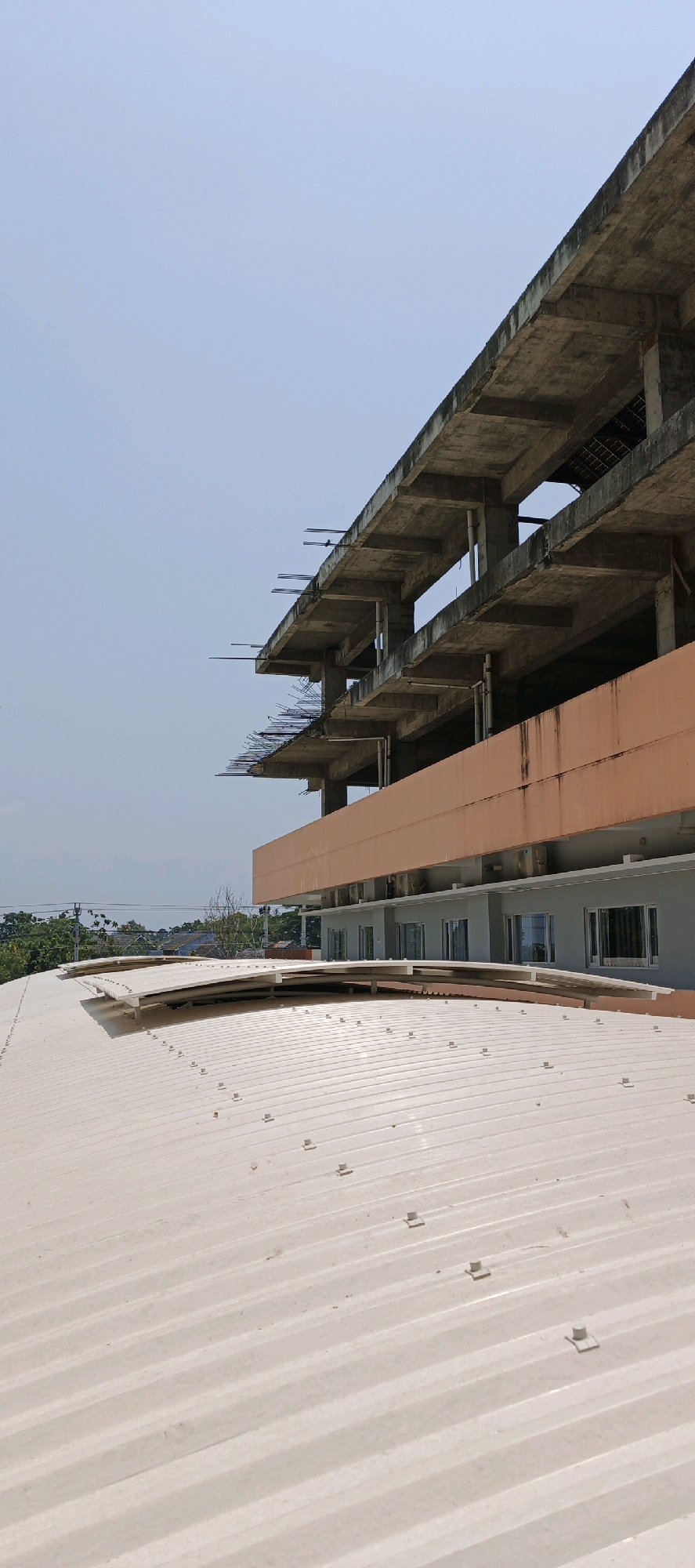 The image, taken in Ambarketawang, Indonesia, depicts an unfinished concrete building with multiple exposed floors and structural elements, contrasting with a lighter-colored corrugated metal roof in the foreground. The sky above is clear and light blue. In the middle ground, a section of the building features an orange-brown facade with several visible windows, suggesting it is a completed part of the structure. Trees and distant buildings can be seen behind the construction site, indicating an urban or semi-urban environment. The foreground is dominated by the metallic roof, characterized by its ridged texture and visible fasteners. There are no people or animals present in the image, and no actions are being depicted. The scene appears to be a static view of a construction project.