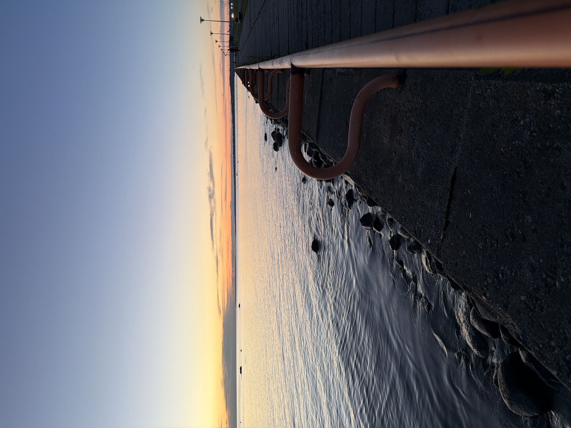 The image depicts a sunset over the Baltic Sea at a pier in Tallinn, Estonia. The sky transitions from a soft blue at the top to warm orange and yellow hues near the horizon, where the sun has set. The sea is calm, with gentle ripples reflecting the sky's colors. The foreground features a concrete pier with a rusty orange railing. The pier extends into the distance, lined with lampposts. Rocks are visible along the edge of the pier where it meets the water. No people are clearly visible, though a distant figure might be present on the pier. The scene conveys a peaceful, end-of-day atmosphere.