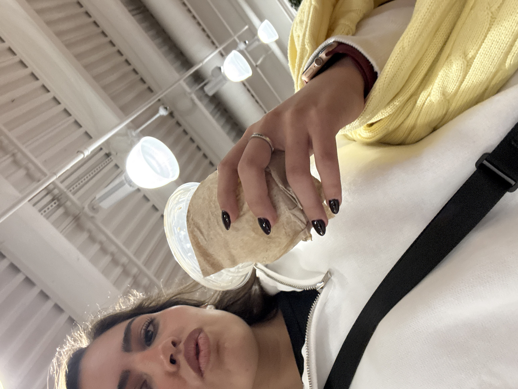 The image captures a person holding a wrapped item, possibly food, in their hand. The person has long brown hair, light skin, and is wearing a white jacket with a black top underneath. A yellow sweater is also visible. The person's hand is adorned with a ring and has black nail polish. A black strap crosses their torso. The background shows industrial-style lighting and a white ceiling.
Without additional context, pinpointing the exact location in San Marcos, United States of America, is impossible. However, based on the ceiling and lighting, it could be a commercial space like a grocery store or a retail outlet.