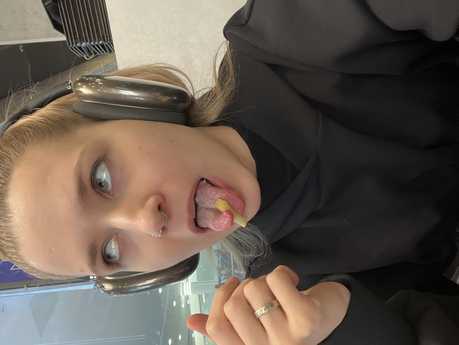 A young Caucasian female is depicted in a close-up, chest-up view, appearing to be in an indoor public setting. She has light-colored hair, a septum nose piercing, and wears large, metallic-gray over-ear headphones, a black hooded sweatshirt over a black undergarment, and a textured silver-colored ring on her right ring finger. Her eyes are rolled upwards, showing only the whites. Her mouth is open with her tongue extended, vertically gripping a single french fry between the tip and a visible fold on the tongue's surface. The background is blurred but reveals a brightly lit interior with a large glass panel on the left, reflecting ambient lights and indistinct shapes. To the right, dark stacking trays are visible. The specific location within the city of Moscow, Russia, is not identifiable from the visual content of the image.