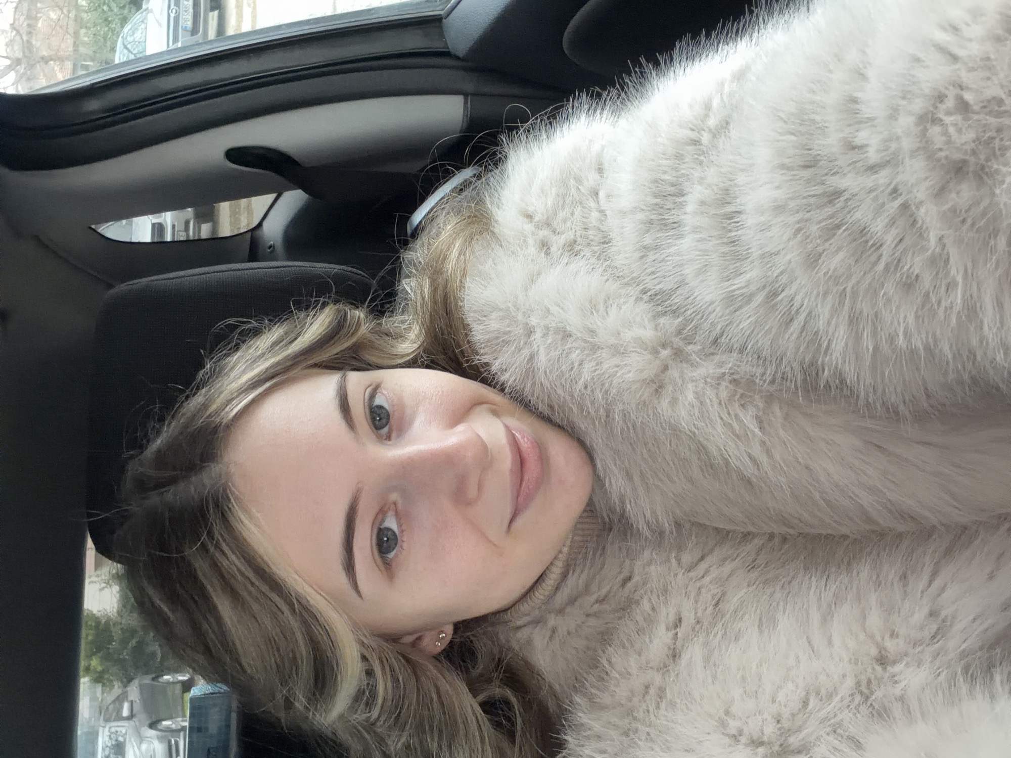 A young woman is posing in a car. She has blonde hair and is wearing a fluffy, light-colored coat. The car's interior is visible, including the headrest of a seat and the car's roof. The location is Plovdiv, Bulgaria, judging from the vehicle's license plate visible in the reflection of the mirror on the roof. Through the car window, other vehicles and trees are visible in the background.