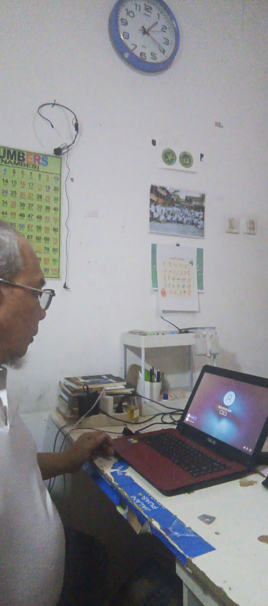The image depicts an indoor scene, likely a desk area in a home or office, with a person operating a laptop. The individual, an adult male with glasses and a beard, is positioned on the left side of the frame, looking towards the laptop. The laptop, which is open and powered on, is on the desk in front of him, displaying a login screen with the text "Administrator."
The desk is cluttered with various items. A stack of books is visible to the left of the laptop. A white organizer with pens and pencils stands behind the laptop, and a small bottle with yellow liquid is also present. Cables are spread across the desk, connected to the laptop and other unseen devices.
The wall behind the desk features several decorative and educational items. A round analog clock with a blue frame is mounted high on the wall. Below the clock, on the left, is a chart with numbers and the word "NUMBERS (NAMBES)". To the right of the number chart, there are two circular Islamic-themed stickers, one with Arabic calligraphy. Further to the right, a framed picture shows a group of people, possibly students, in an outdoor setting. Another document with colorful text and images is mounted below the framed picture.
The overall environment suggests a workspace where study or administrative tasks might be performed. The specific location in Sukaresmi, Indonesia is not discernible from the visual elements of the image itself, but the presence of Islamic decorations could indicate a cultural context common in Indonesia. The lighting appears to be artificial, likely from a room light or the laptop screen.