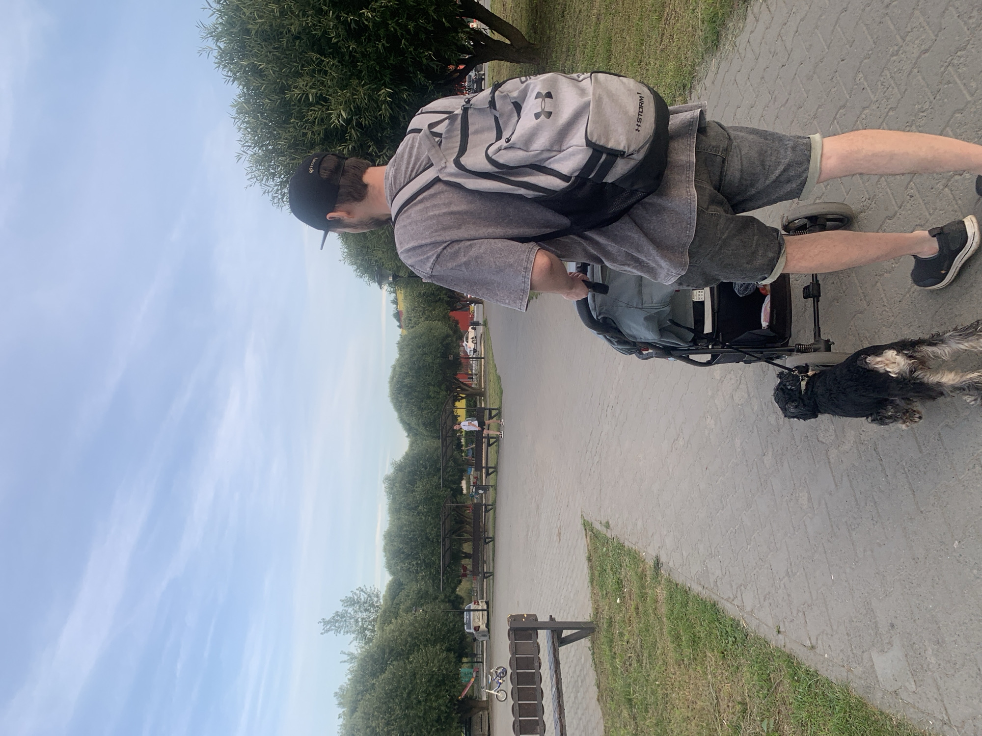 In Chaikovsky, Russia, a man wearing a grey t-shirt, denim shorts, and a black baseball cap pushes a stroller down a paved walkway. He carries a grey and black backpack. A small black dog on a leash walks beside the stroller. The background features a grassy area, benches, trees, and a children's playground with a bicycle. The sky is partly cloudy.