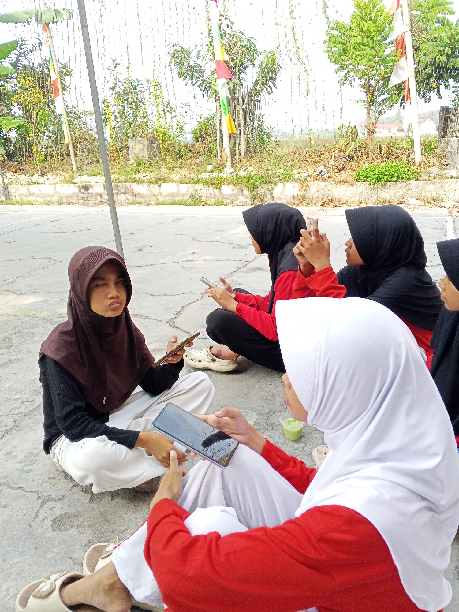 The image depicts five individuals, four of whom are visible and appear to be young women, wearing hijabs and modest clothing. They are seated on a paved outdoor area, possibly a sports court or schoolyard, in what is identified as the city of Ciruas, Indonesia. The environment includes vegetation, a fence, and flags, suggesting an outdoor event or gathering. Two individuals are holding smartphones, one of whom is taking a picture or video. Another individual is looking at their phone, and the person in the foreground with a white hijab is also engaged with a phone. The overall scene suggests a casual gathering where the participants are interacting with their mobile devices.